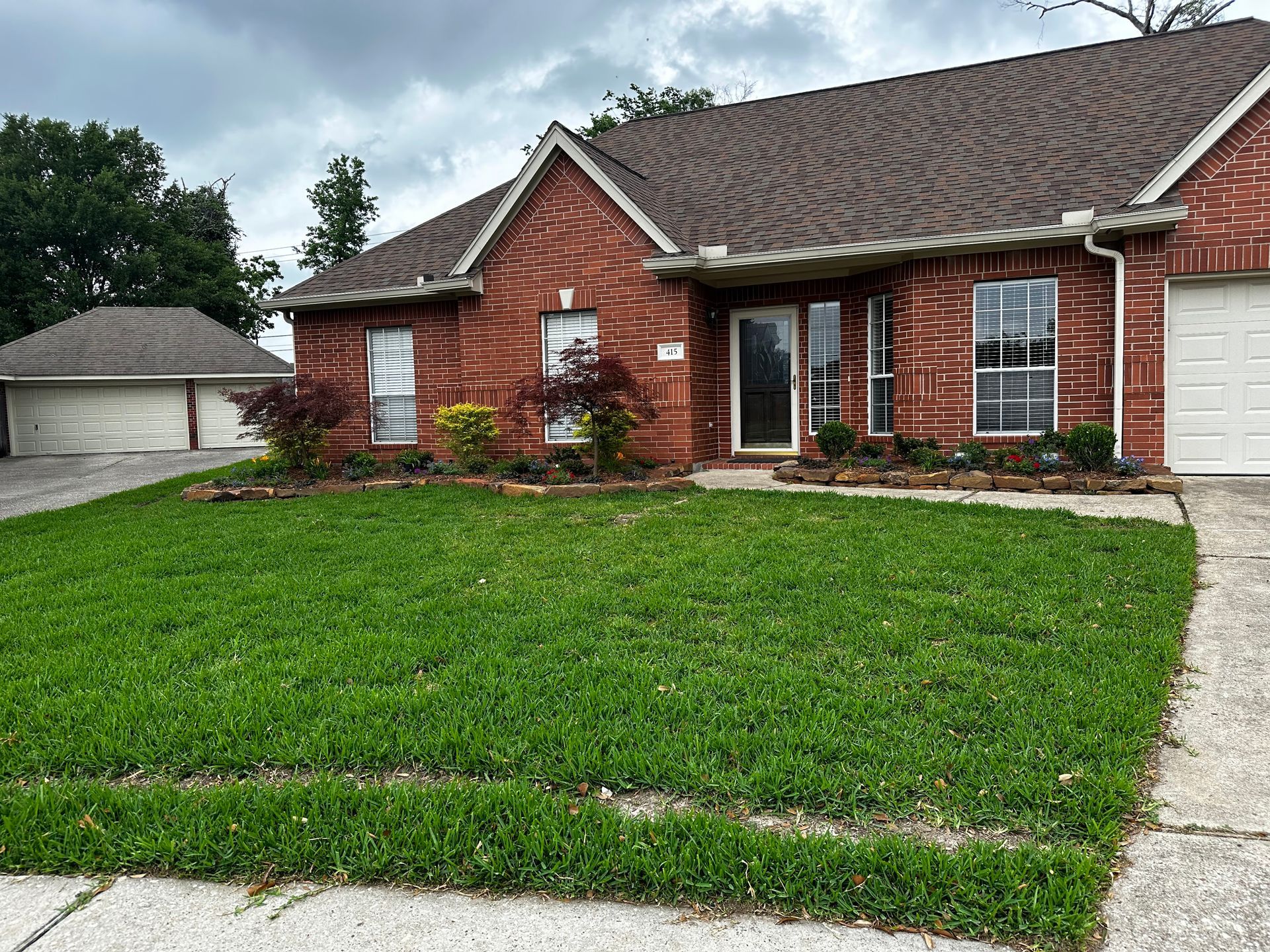 Red brick house with green lawn, flowerbeds, and garage on a cloudy day.