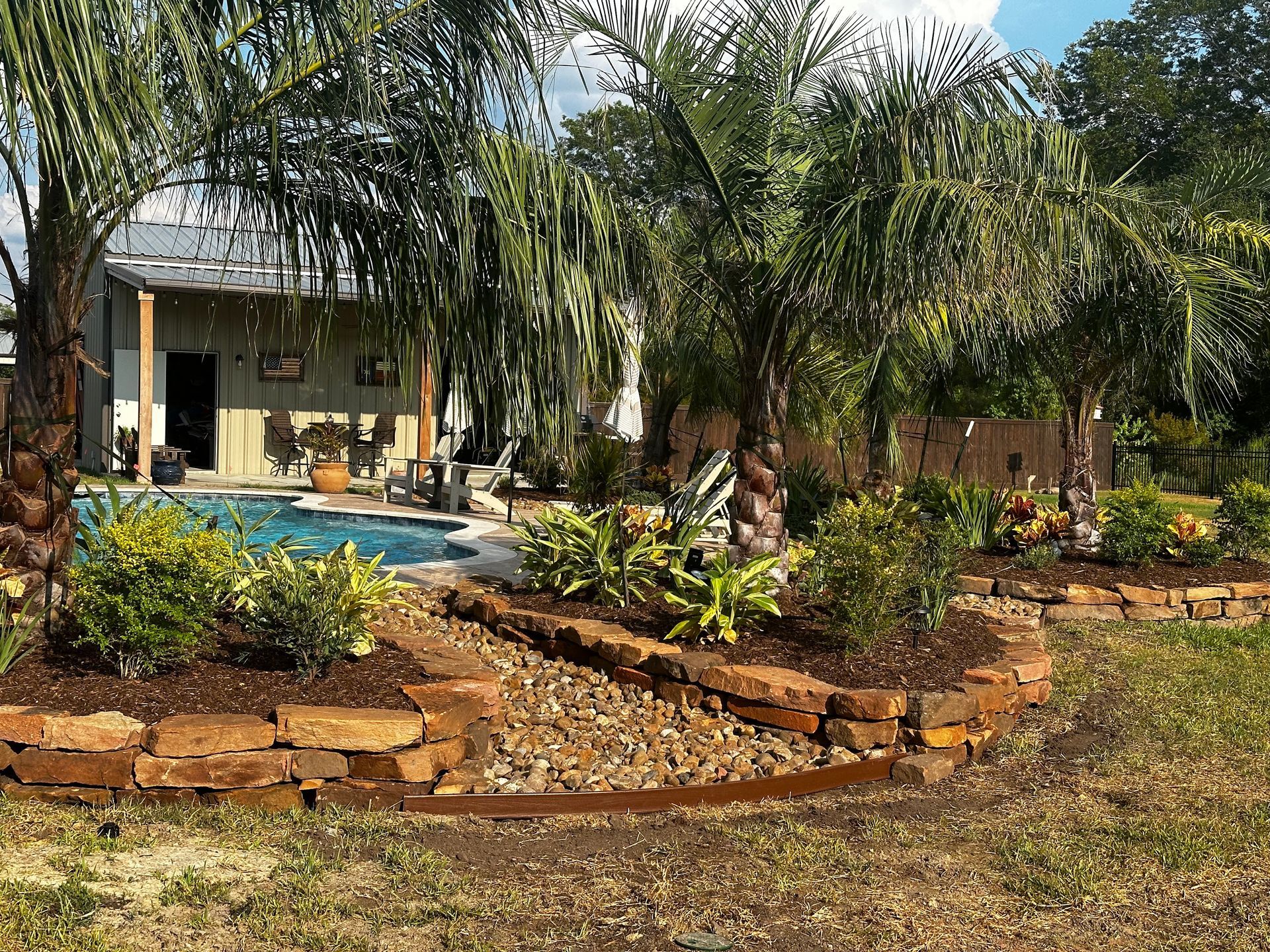 Landscaped backyard with pool and stone retaining walls. Lush greenery and palm trees surround the water.
