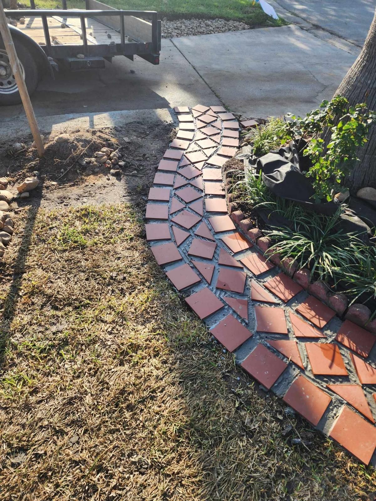 Brick pathway curves through a yard, surrounded by grass and a small garden near a tree.