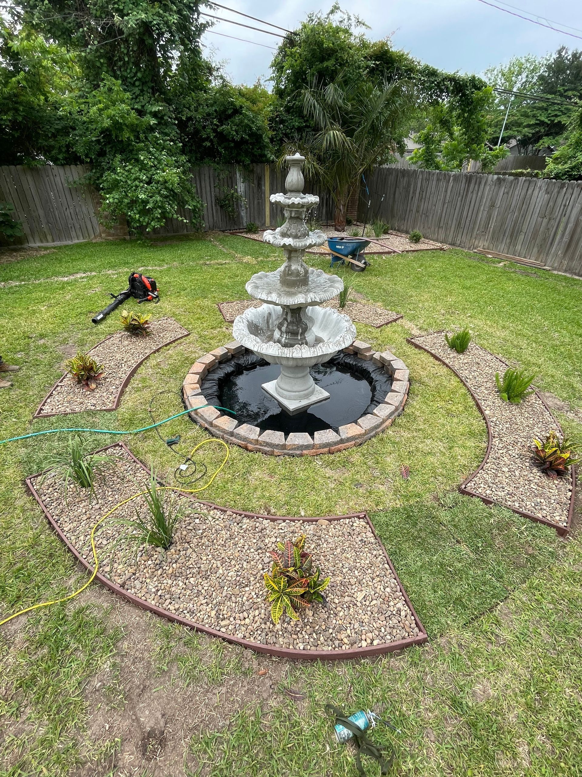A backyard with a tiered fountain as the central focus, surrounded by a grassy area and rock beds.