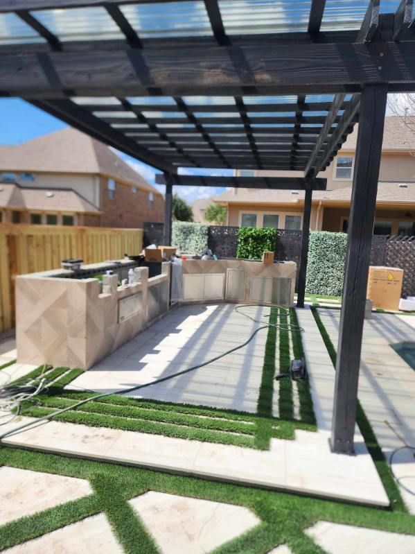 Outdoor kitchen under a pergola with green turf accents; stone counters and cabinets; sunny day.