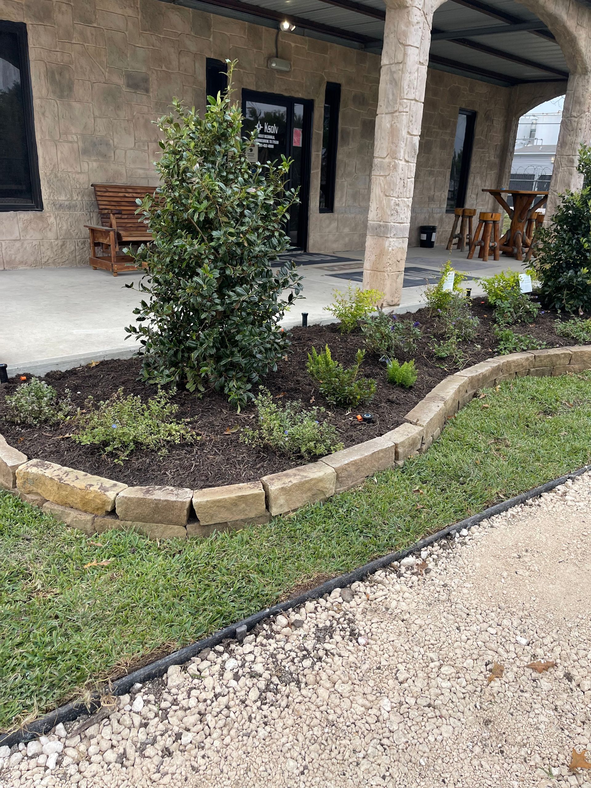 A building with a stone facade and arched columns. A raised garden bed with plants in front.