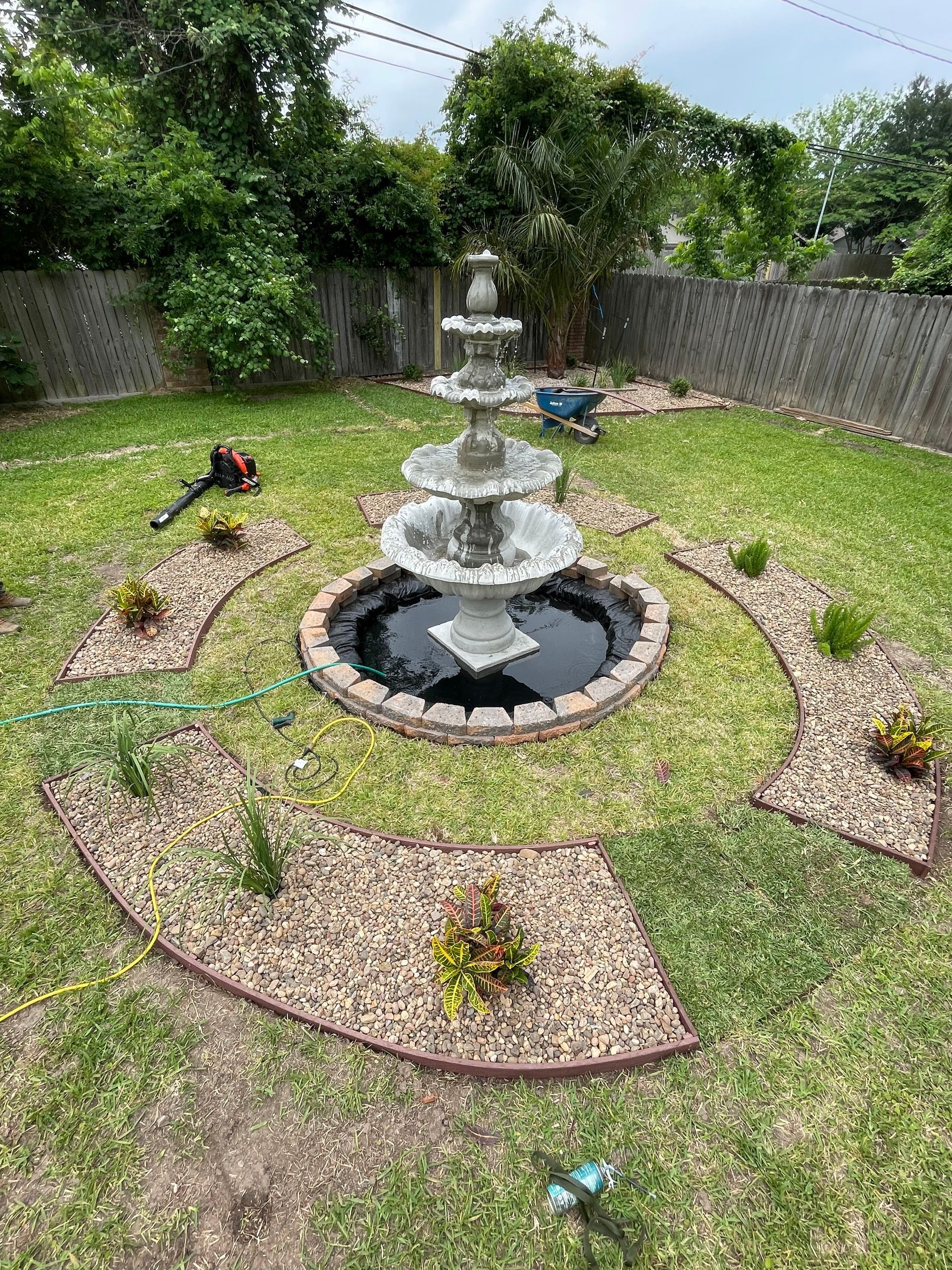 A tiered fountain in a grassy yard with decorative rock beds and surrounding greenery.