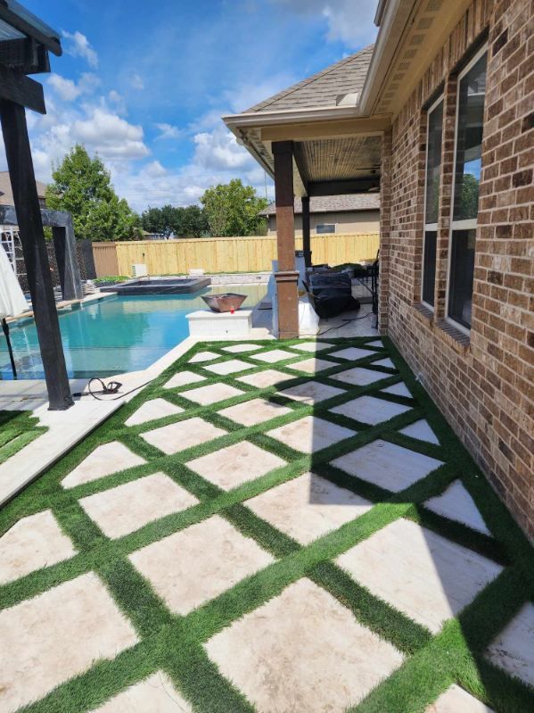 Patio with diamond-patterned artificial turf, concrete pavers, pool, and brick house. Bright sunny day.