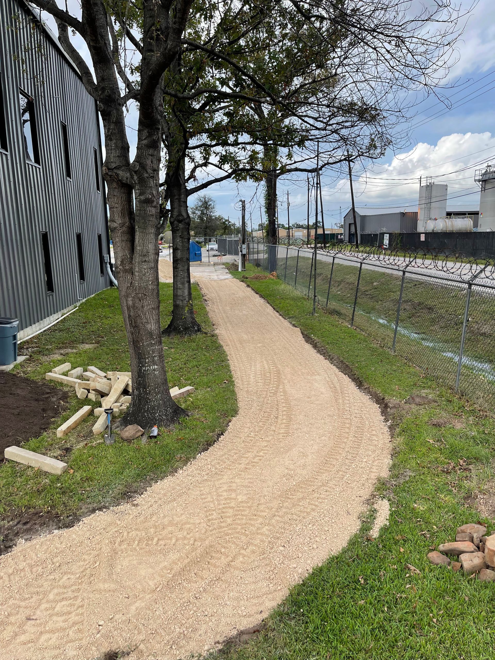 A gravel path winds through a grassy area, flanked by trees and a chain-link fence, leading towards buildings.