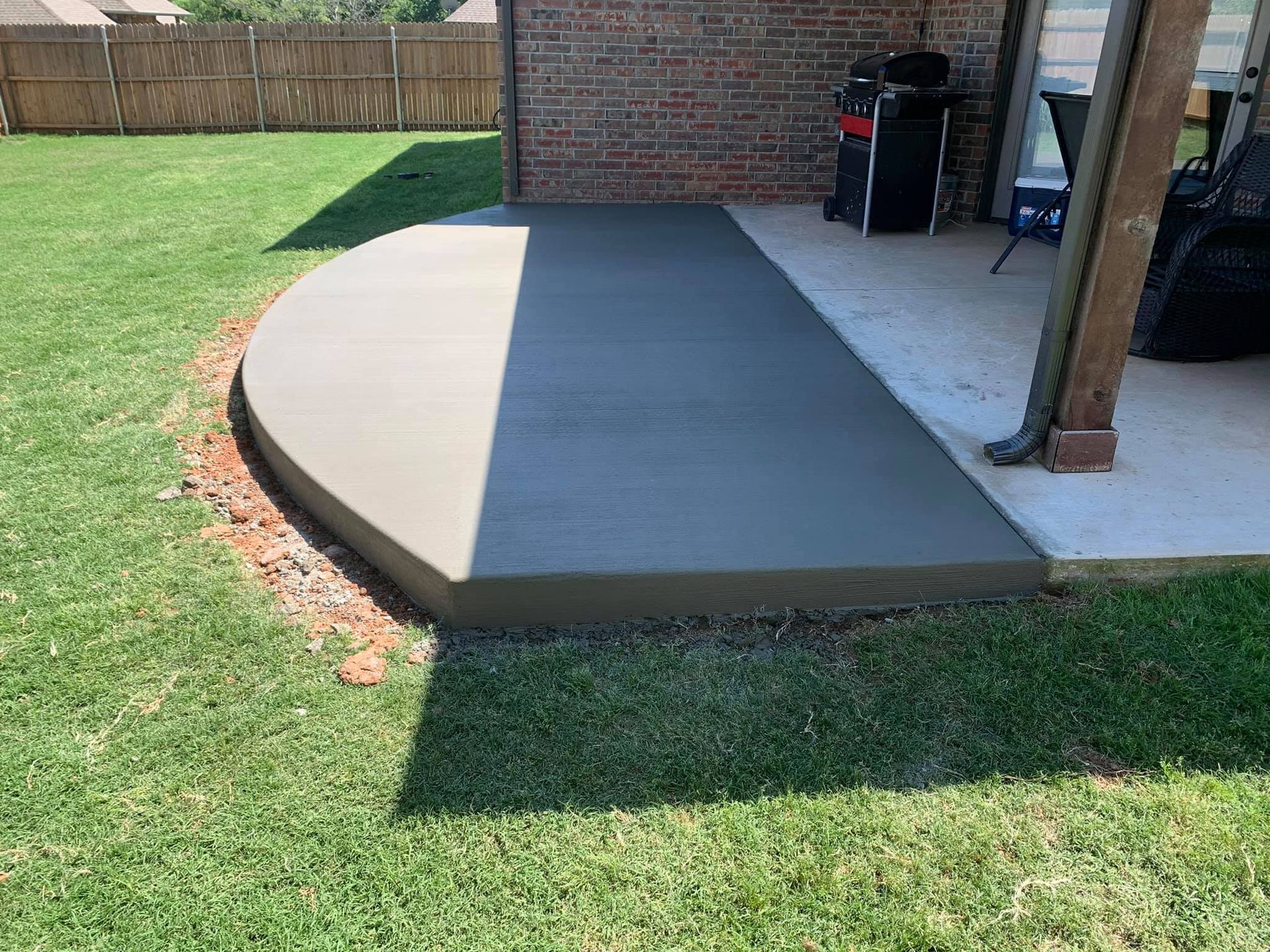 Newly poured concrete patio with curved edge, next to grassy yard and brick wall.