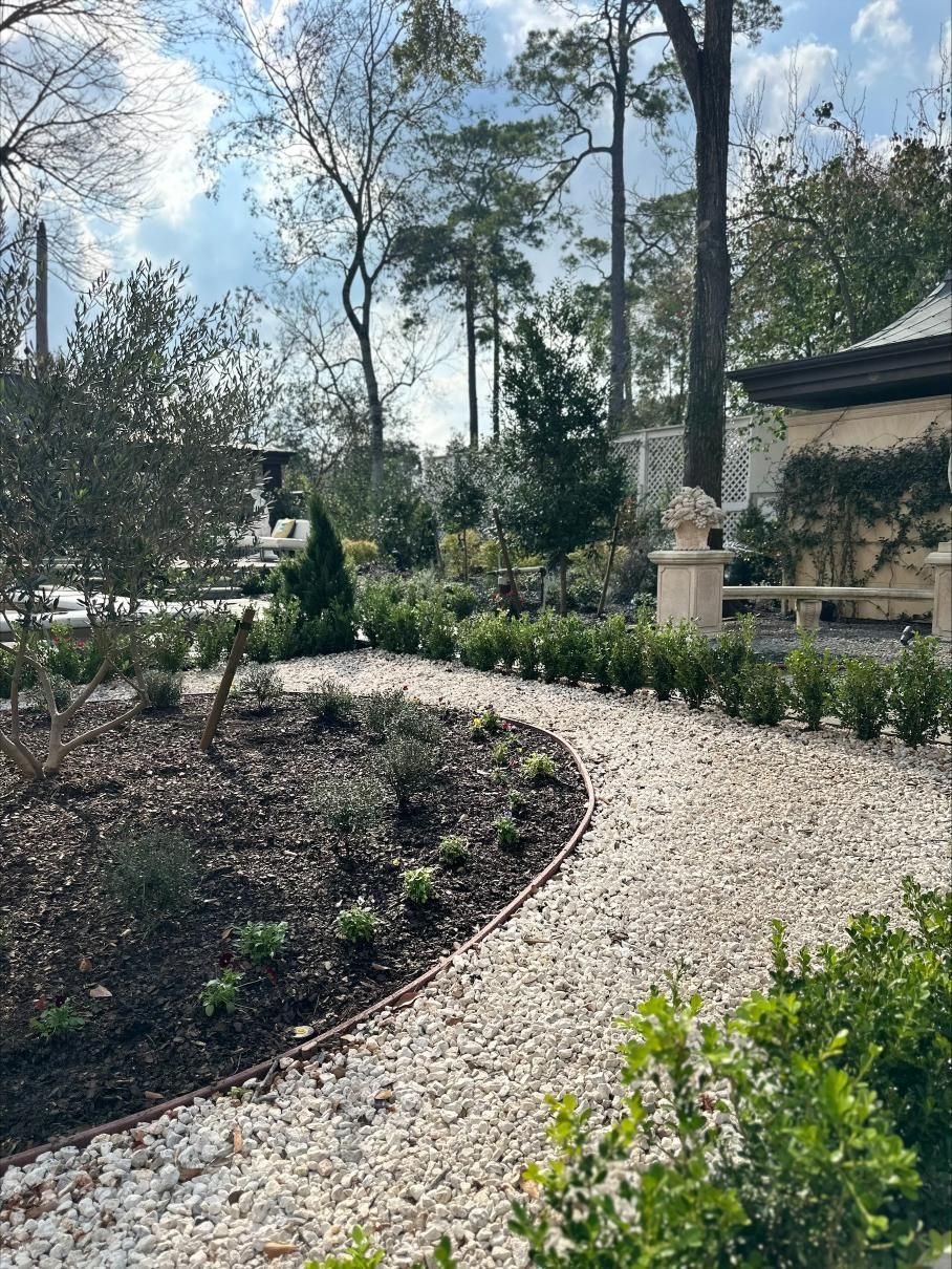 House with beige facade, black roof, surrounded by green lawn, colorful flowers, and trees under a cloudy sky.