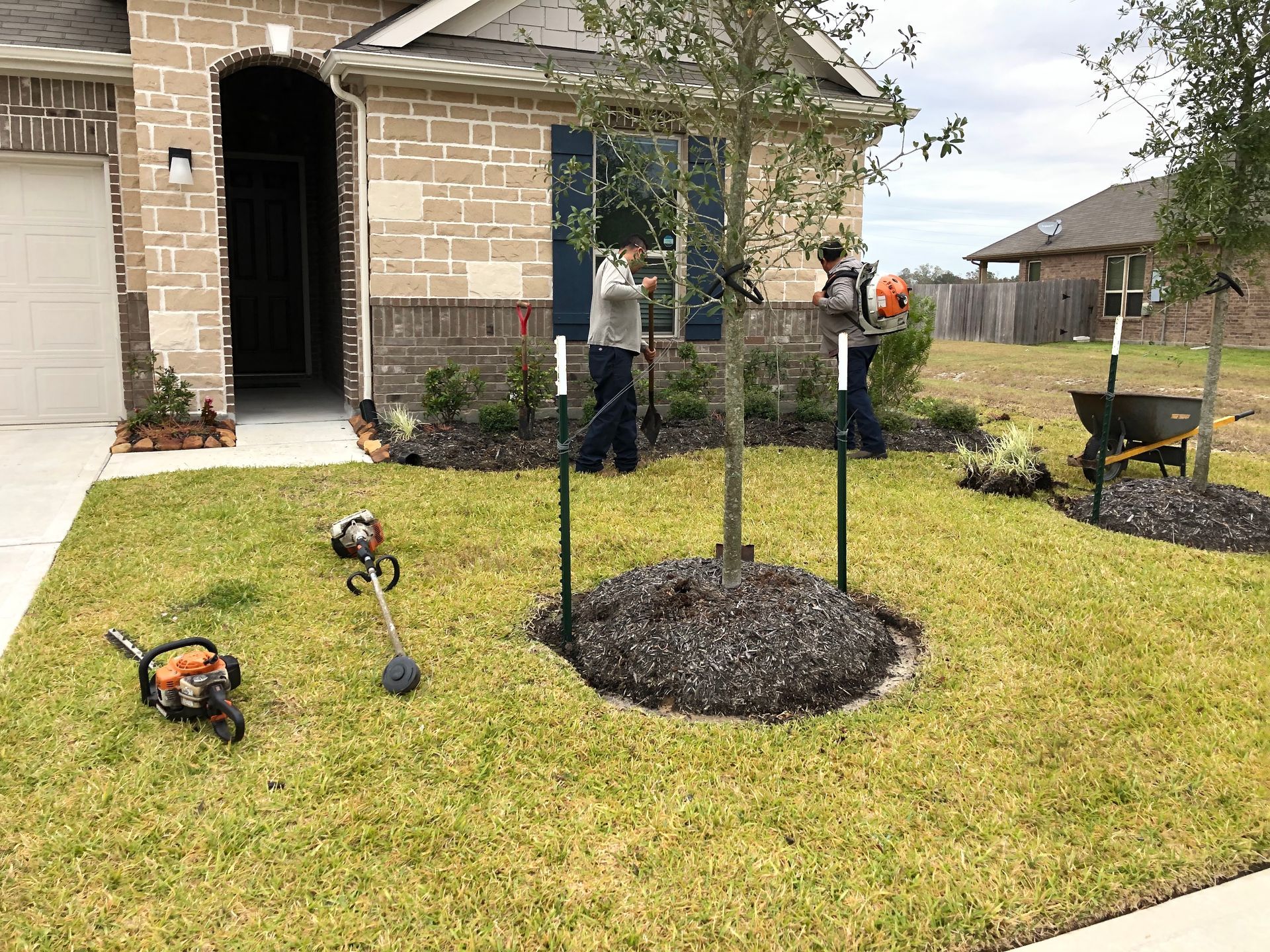 Two people landscaping a yard with a tree and equipment in front of a house.