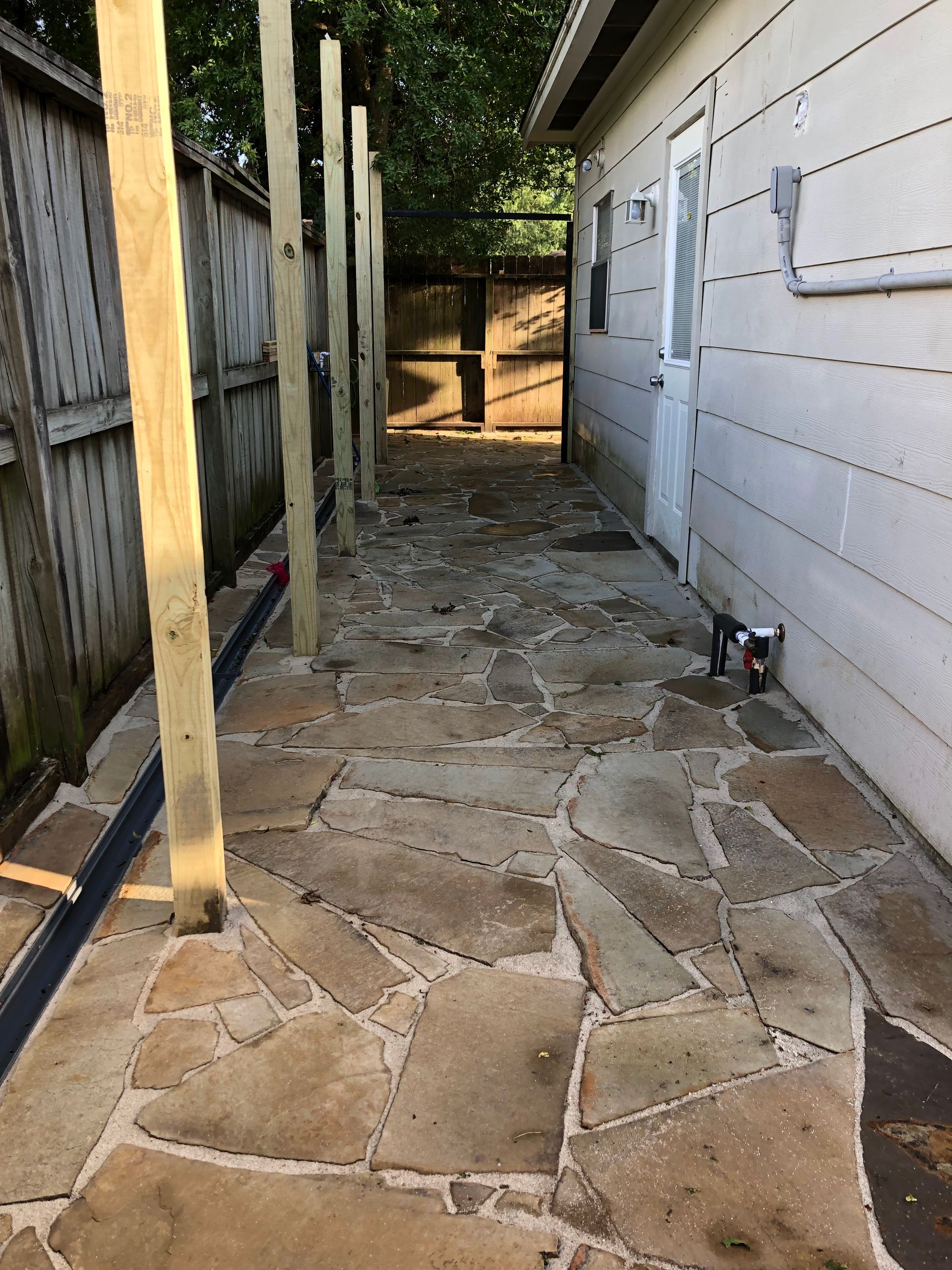 A narrow flagstone walkway with wooden posts alongside a fence and building.