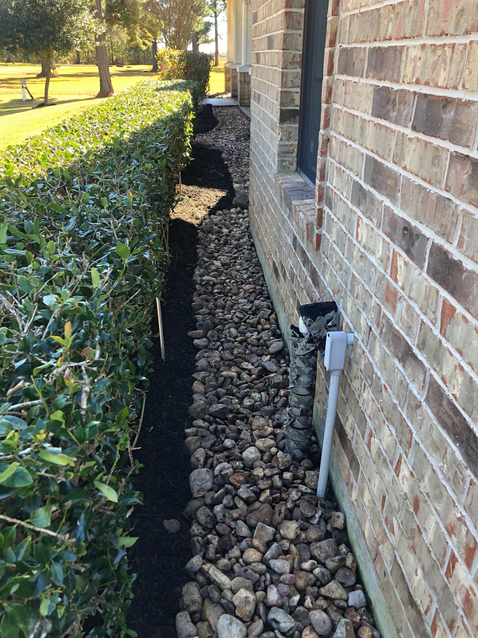 Gravel-filled landscaping bed beside a brick house, next to a green hedge, with a downspout and electrical outlet.