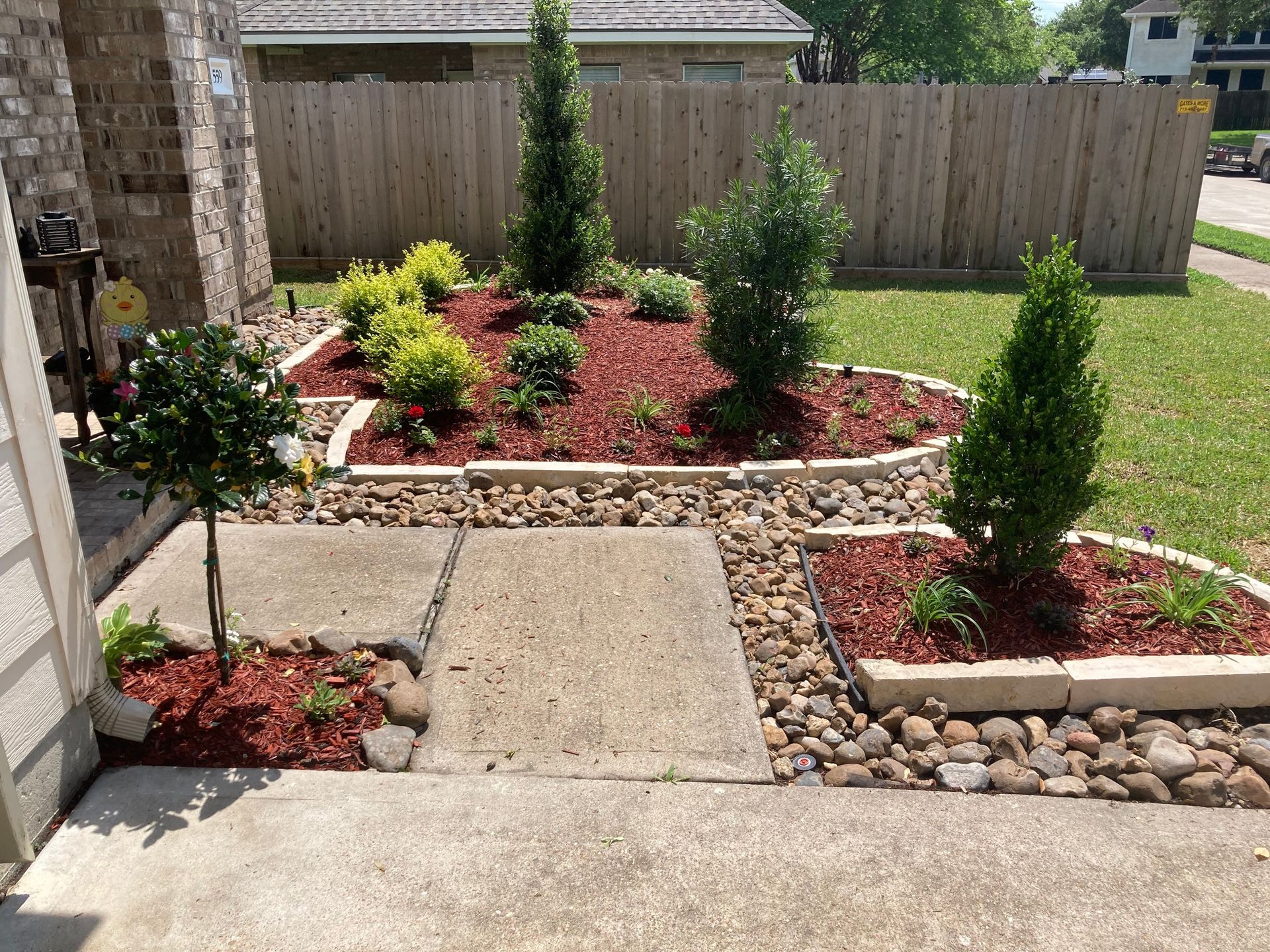 Landscaped front yard with red mulch, stone borders, and various green plants. Concrete walkway.