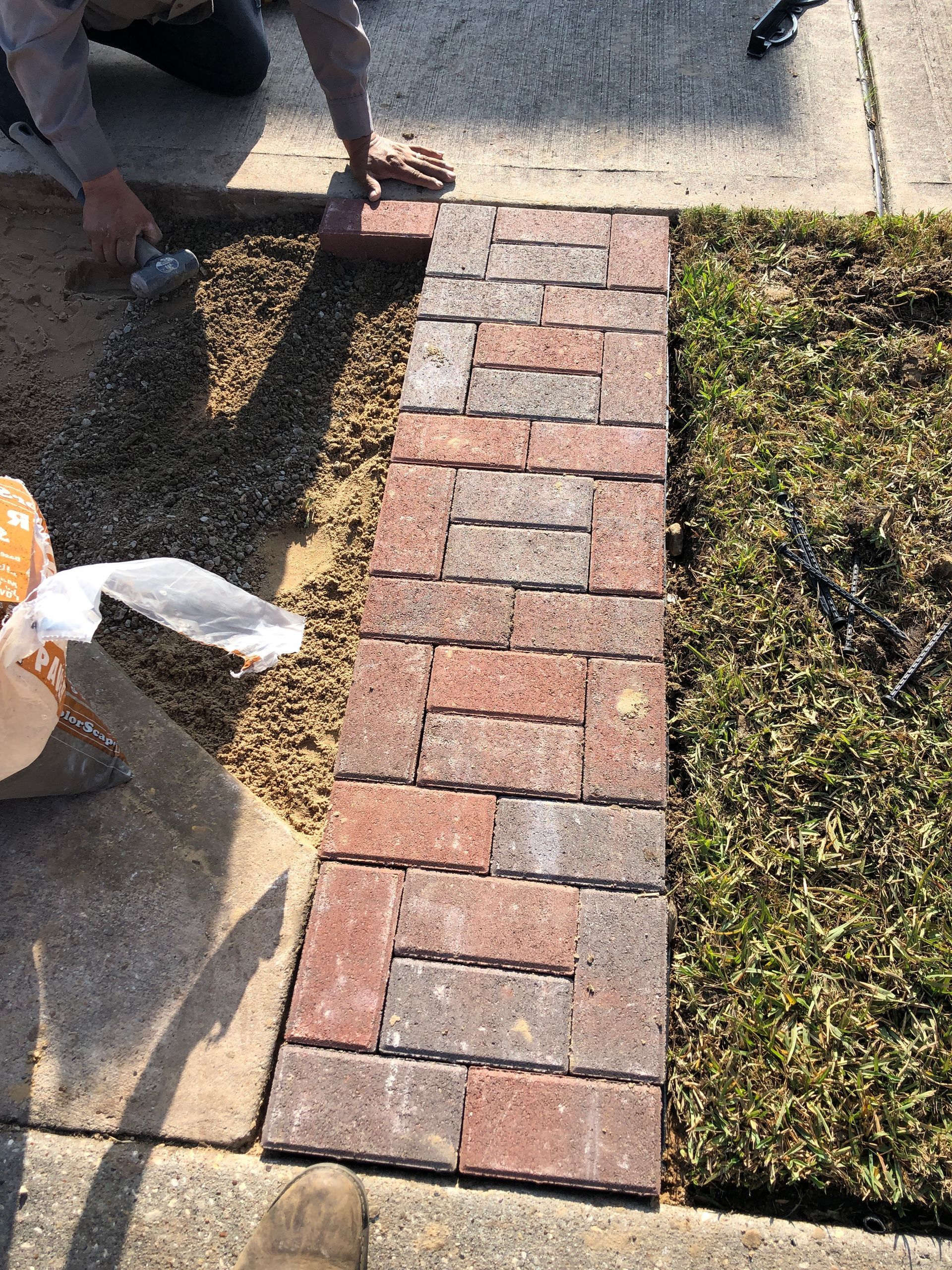 Person laying brick pavers for a pathway next to a sidewalk and grass.