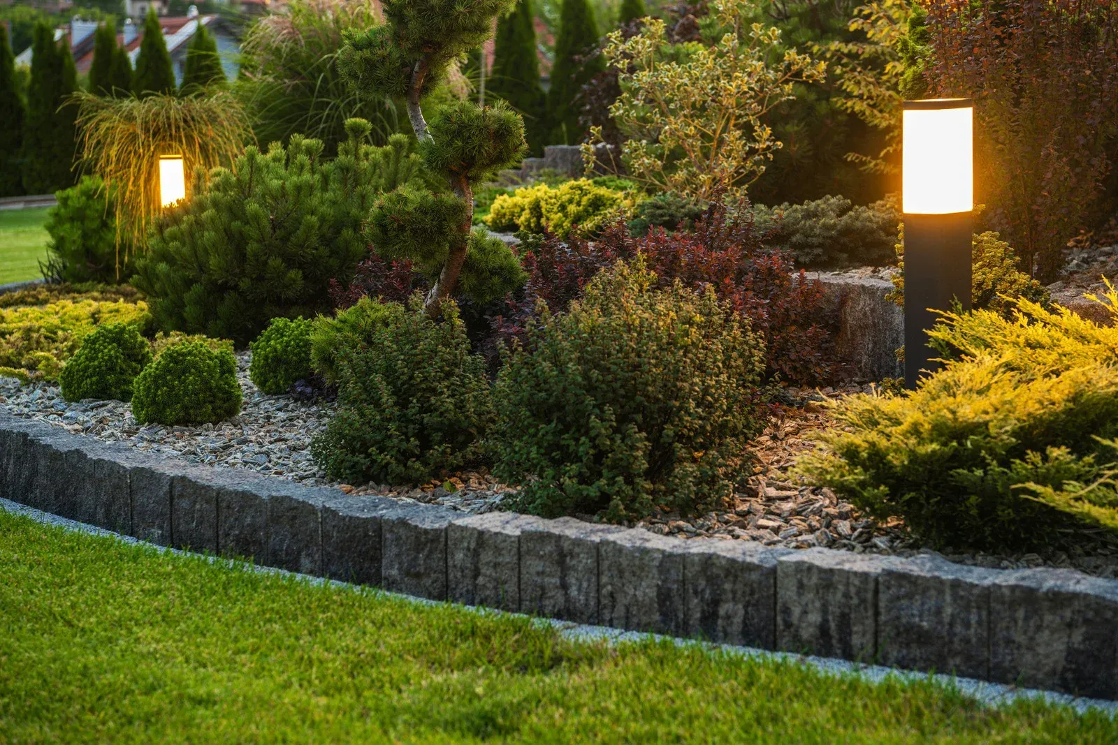 Lush garden bed with various green and red bushes, bordered by stones and lit by modern lamps.