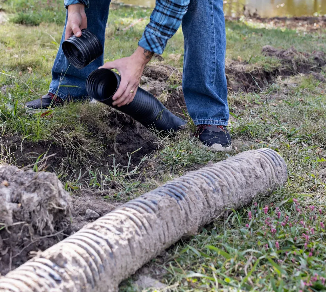 Person connecting black corrugated drain pipes in a grassy ditch.