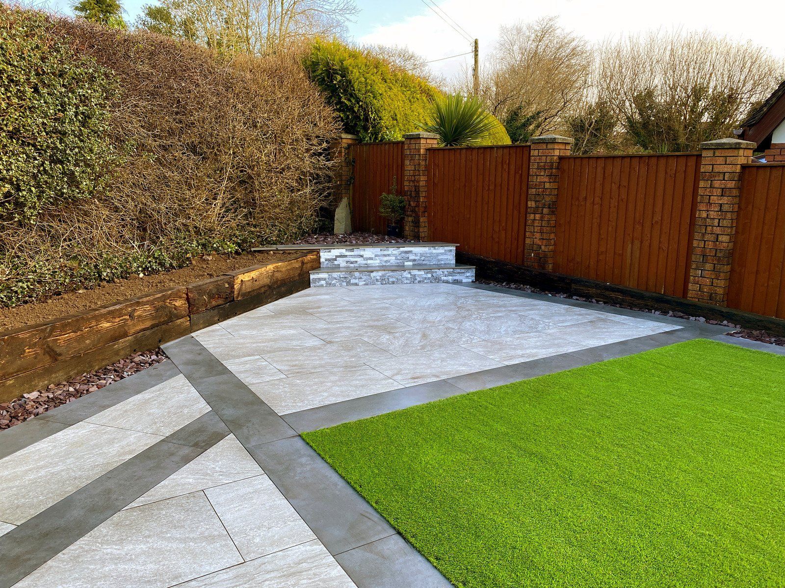 Patio with gray pavers, green turf, brown fence and hedge in a yard.