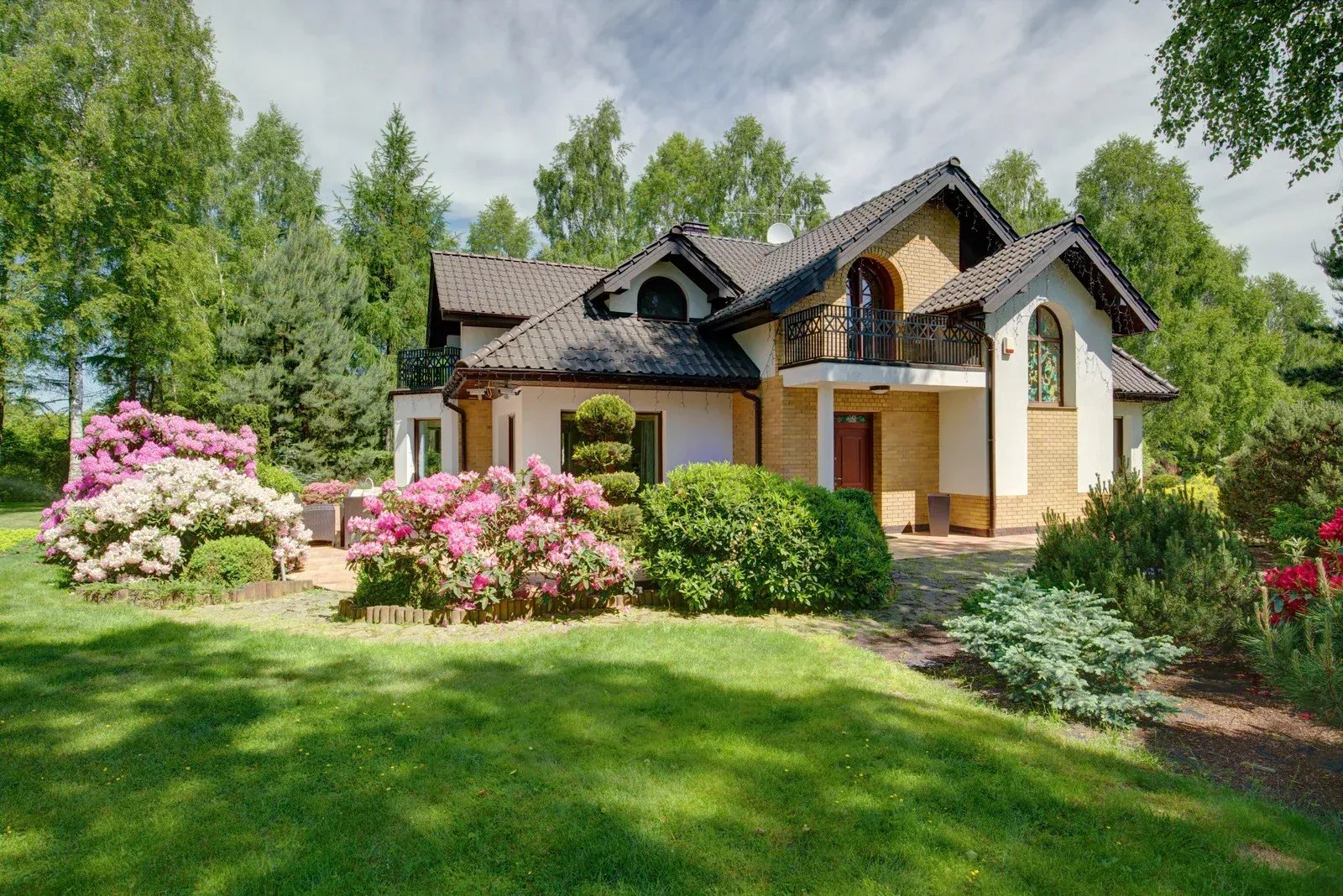 House with beige facade, black roof, surrounded by green lawn, colorful flowers, and trees under a cloudy sky.