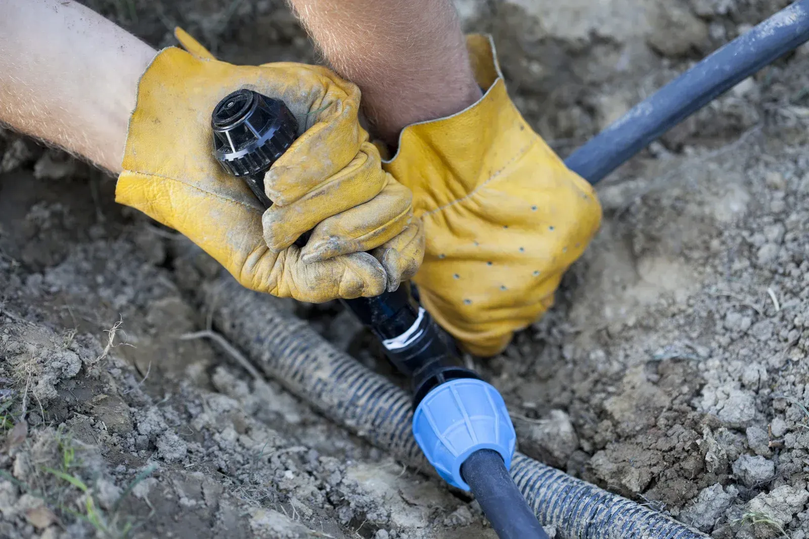 Hands in yellow gloves connecting black plastic pipe to corrugated tubing in dirt.