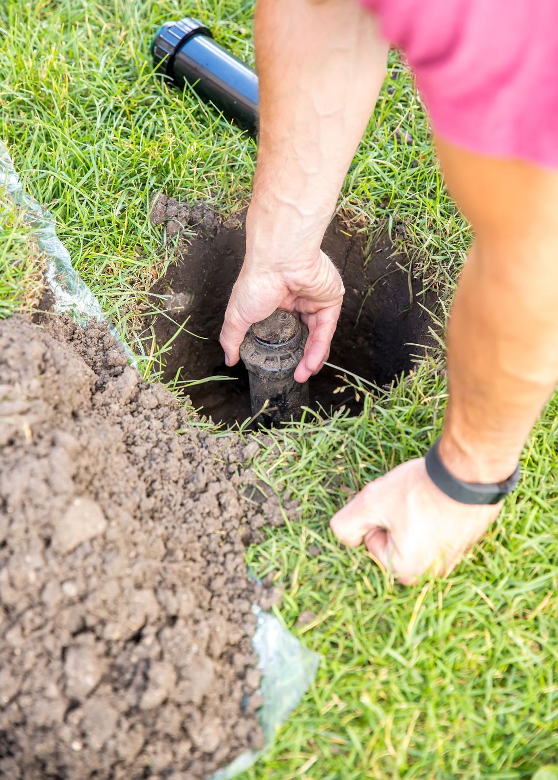 Person installing a sprinkler head in a grassy yard, hands in a dark hole, soil beside.