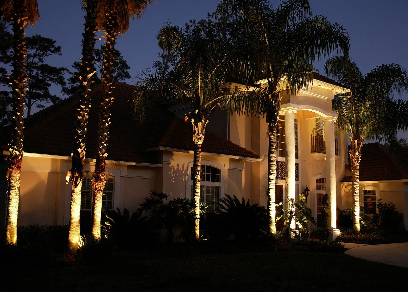 Night view of a large house illuminated by spotlights, showcasing palm trees and architecture.