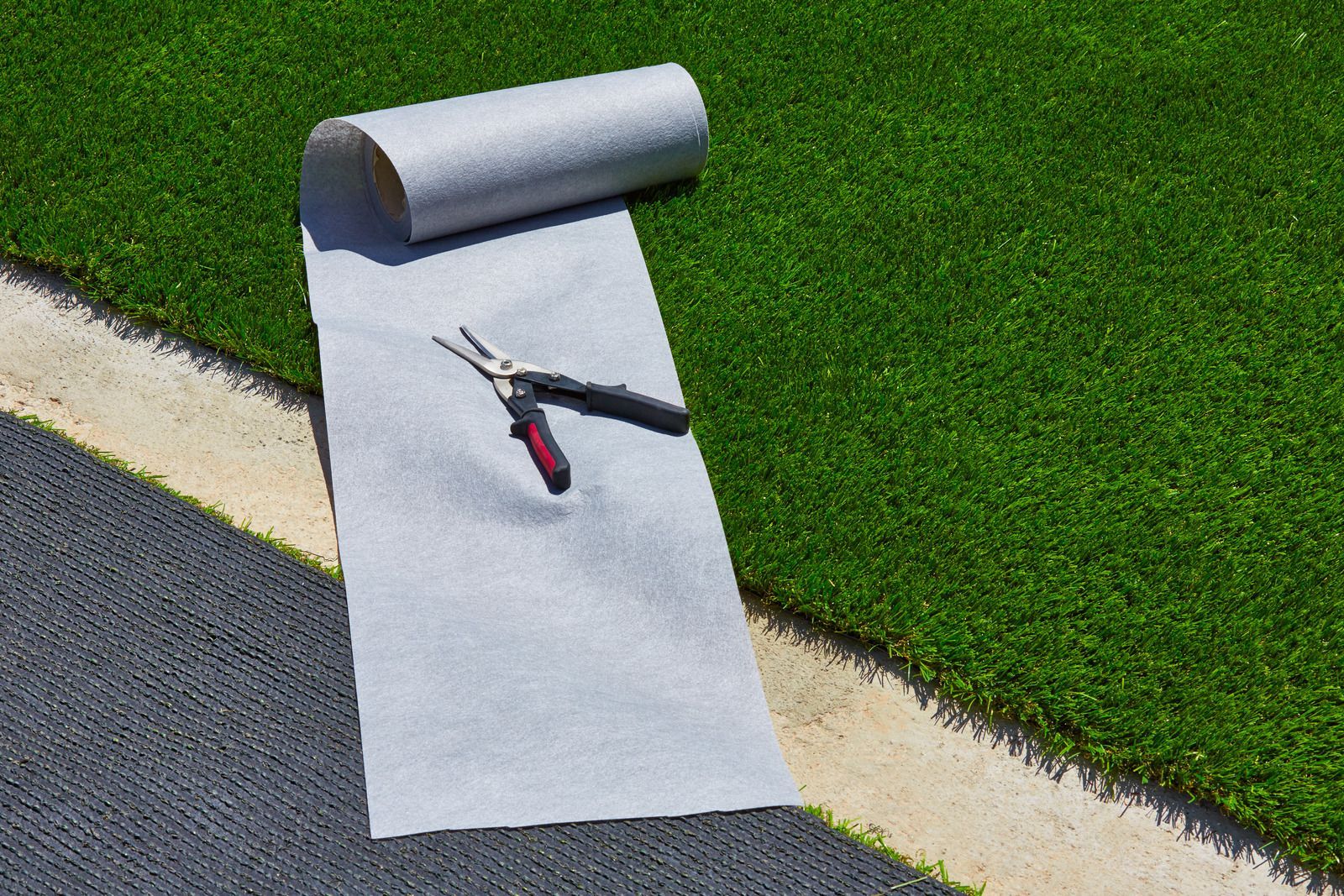 Roll of gray fabric, scissors, and artificial turf being installed alongside a contrasting gray surface.