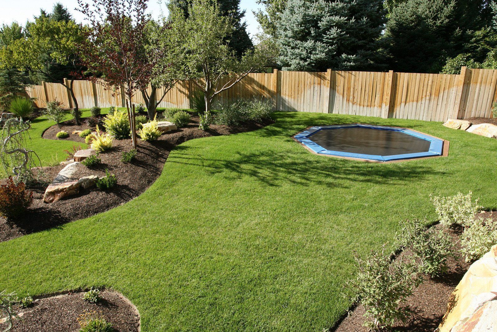 Well-manicured backyard with lush green grass, landscaping, and a trampoline against a bamboo fence.
