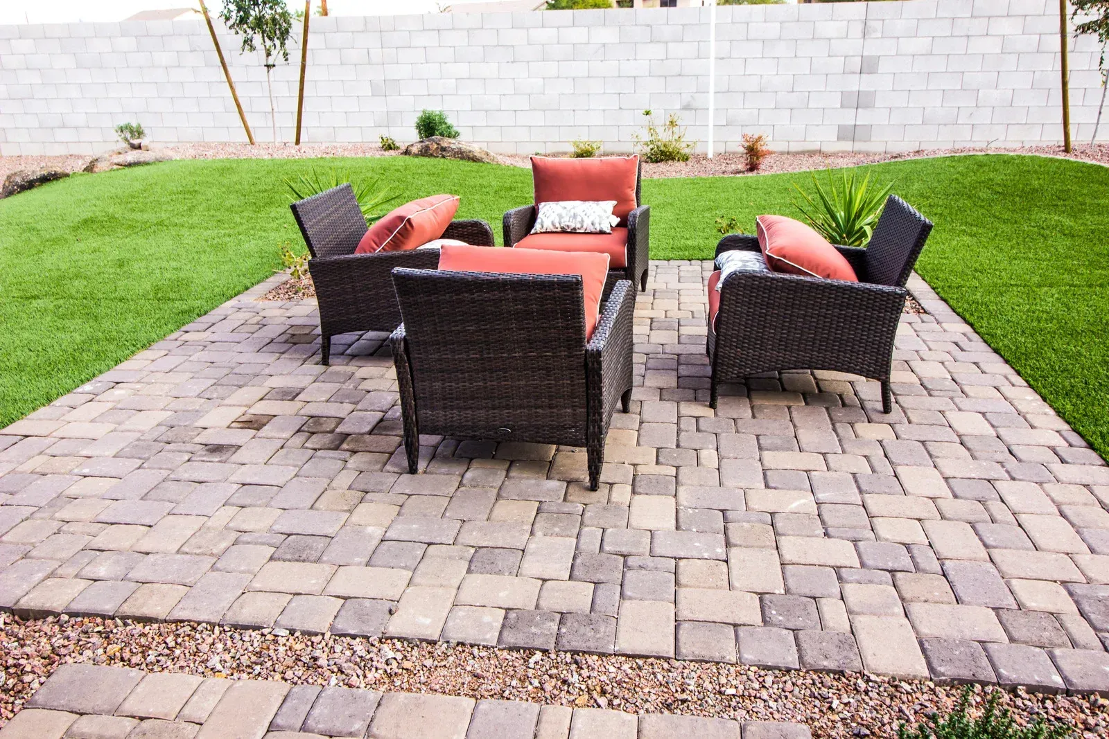Patio with four wicker chairs, orange cushions, on brick pavers; green lawn in background.