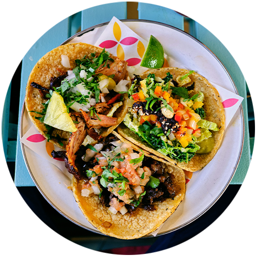 Three tacos are on a white plate on a table