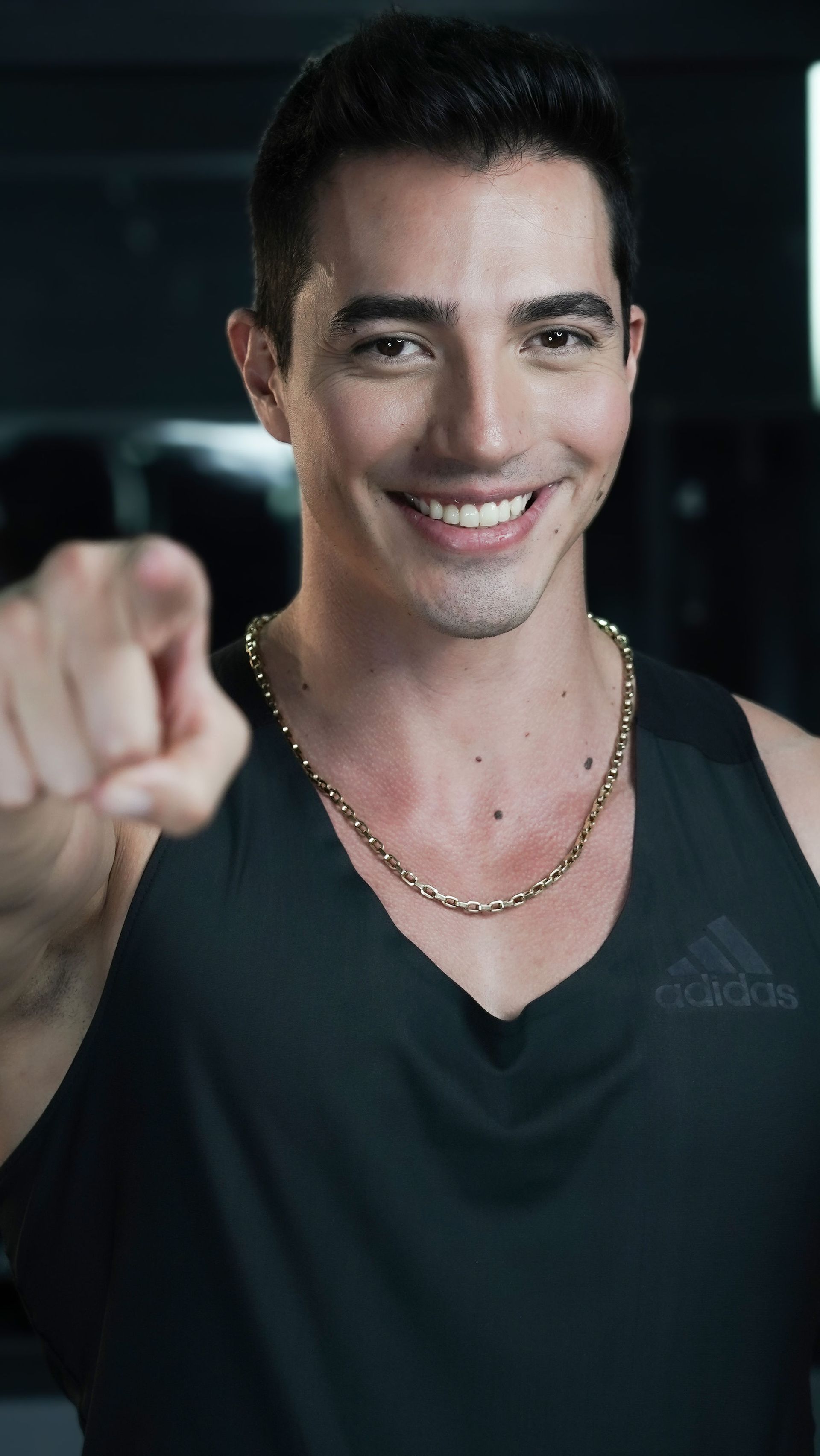 A man wearing a black tank top and a gold chain is pointing at the camera.