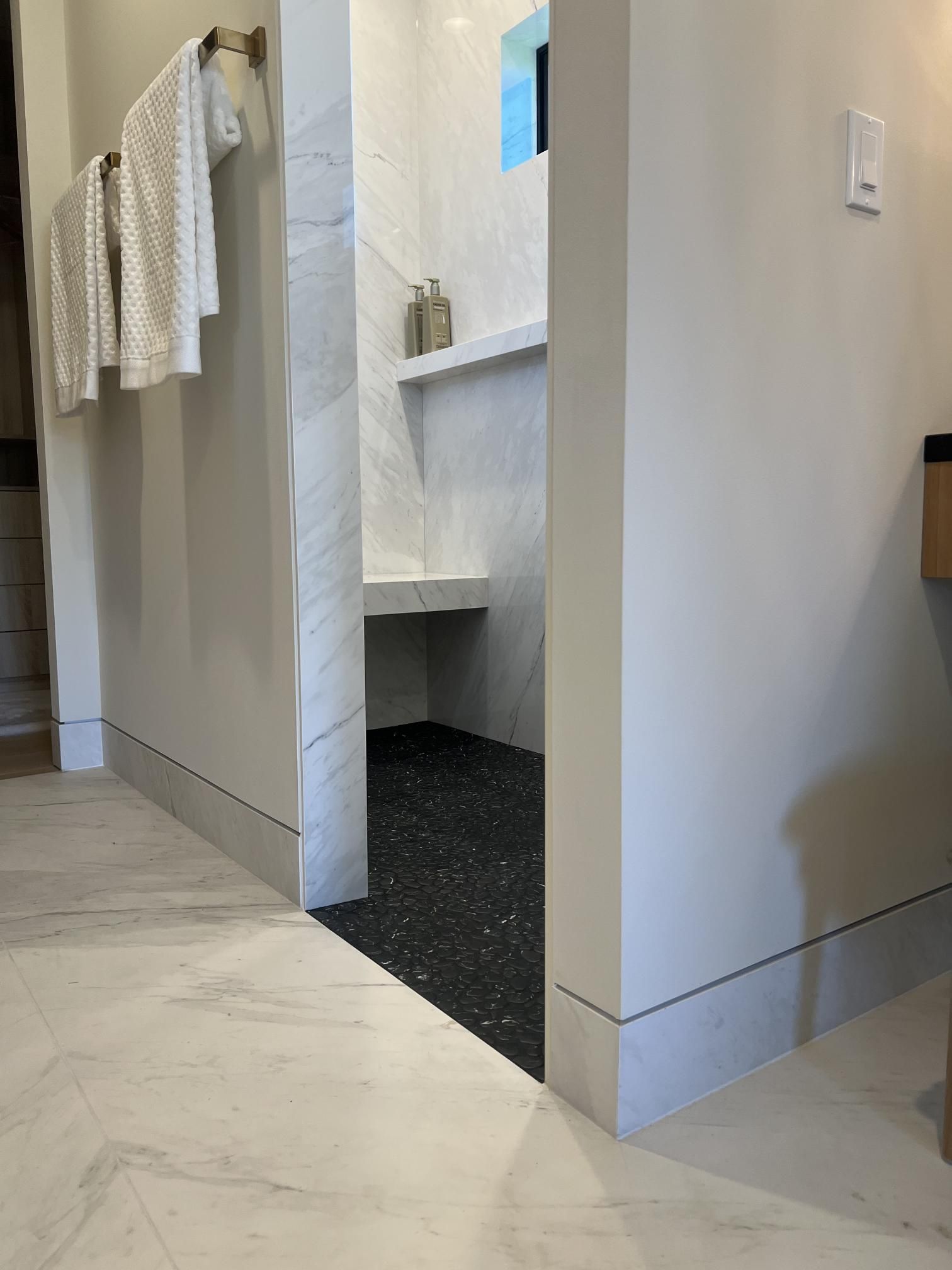 Bathroom with shower entrance. White walls, marble floor, and dark tile shower floor.