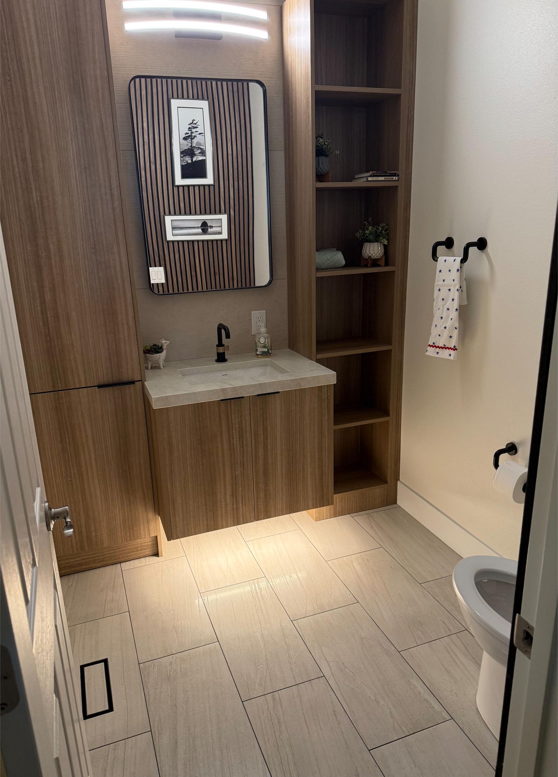 Small bathroom with wooden vanity, mirror, shelving, and white toilet.