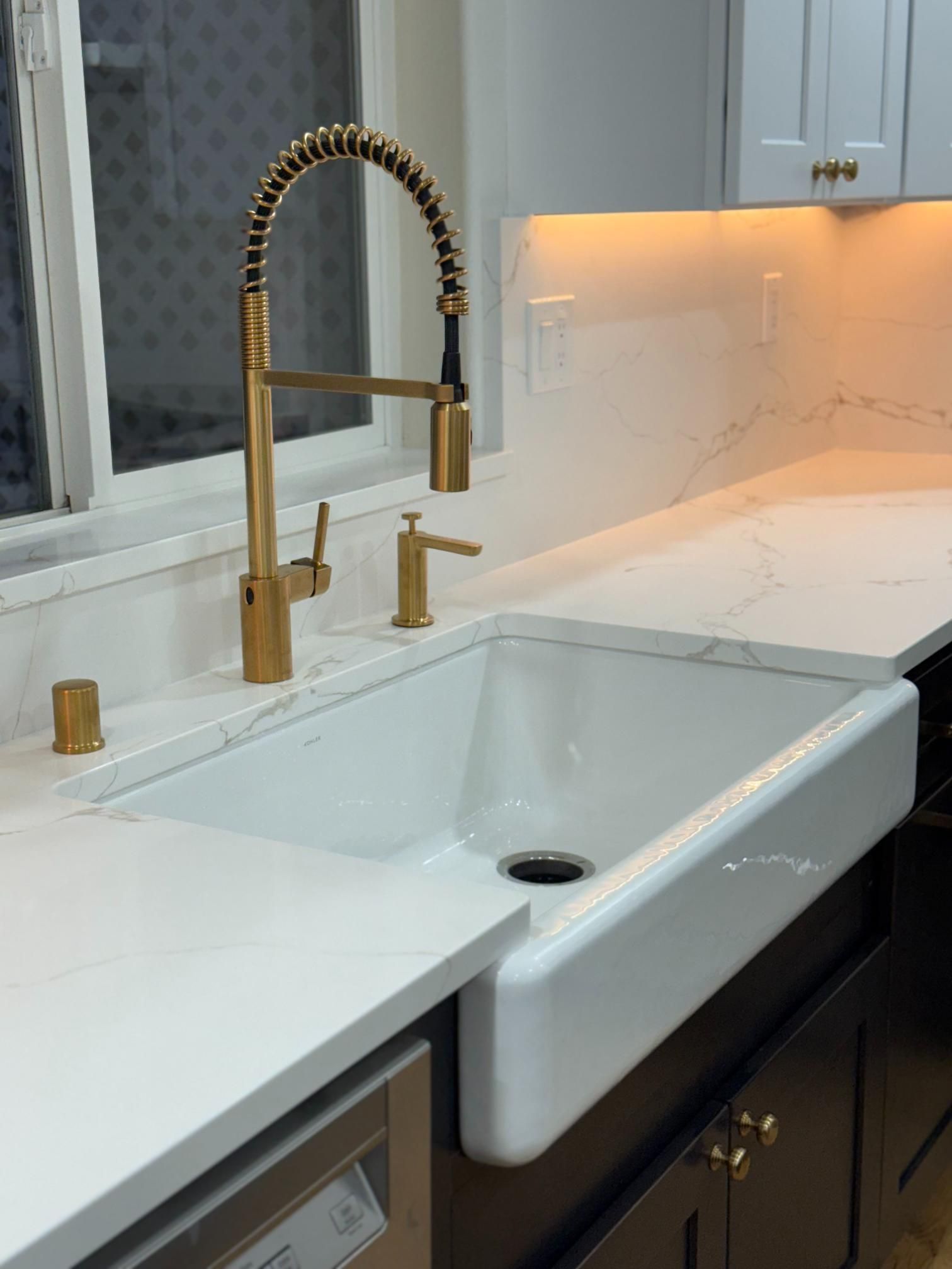 Gold faucet and farmhouse sink in a modern kitchen with white countertops.