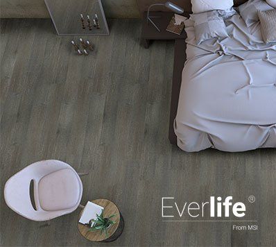 Bedroom with gray wood-look floor, bed with gray bedding, small table, chair, and decor; Everlife logo.