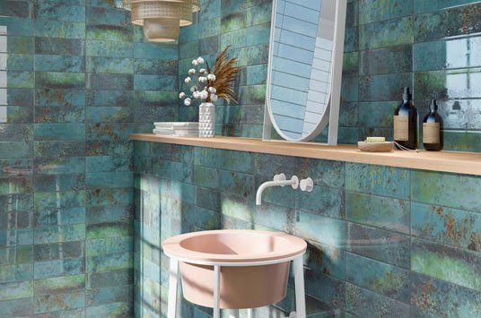 Bathroom with teal and copper-colored brick tiles, pink sink, wooden shelf, and mirror.