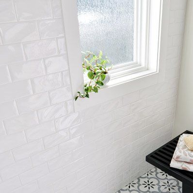 White-tiled bathroom with a window, plant, and bench. Gray and white patterned floor.
