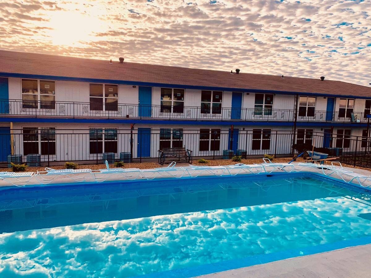 Motel exterior with blue trim and a rectangular pool; cloudy sky with sunlight.