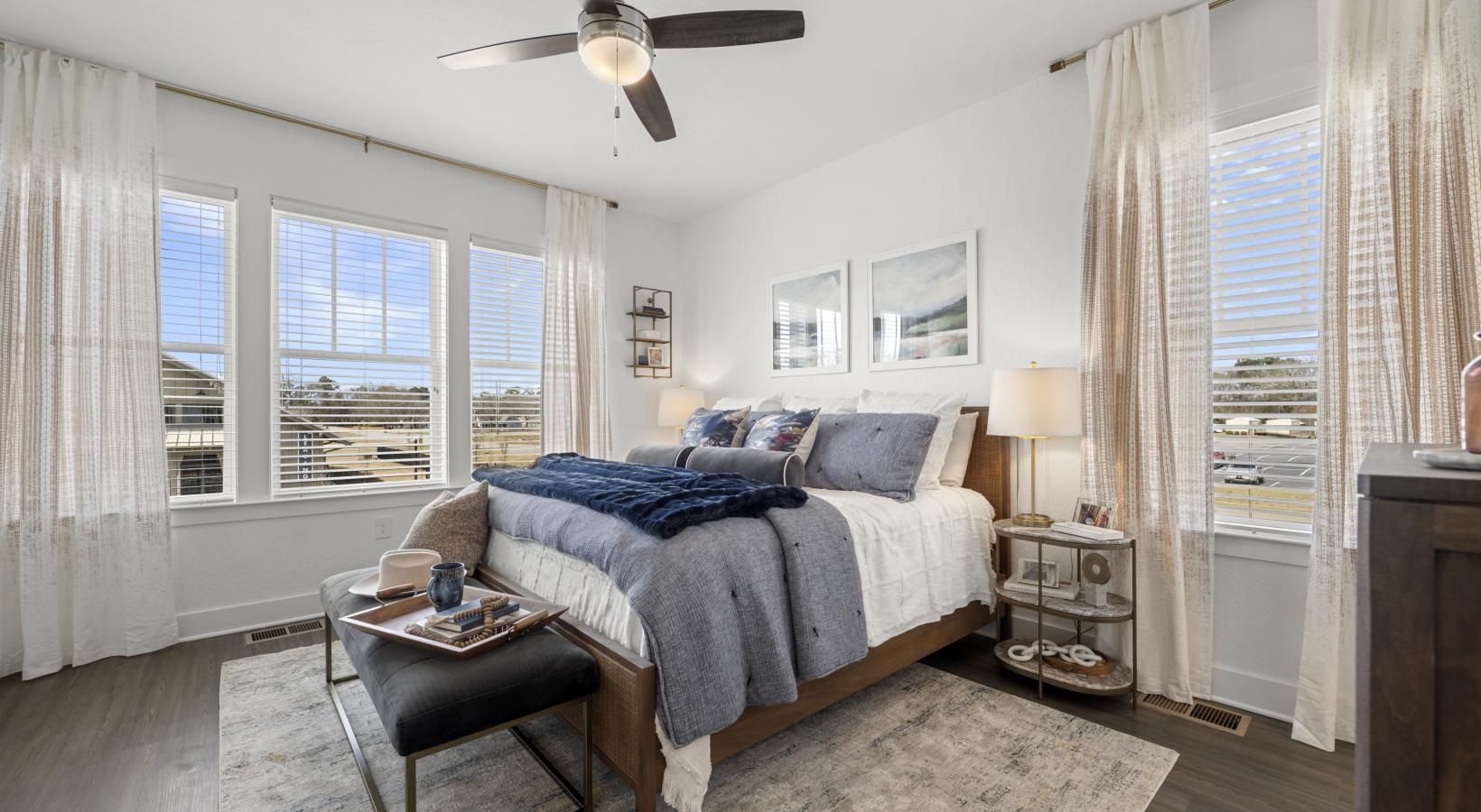 Bright bedroom with a queen bed, bench at foot, large windows, and sheer curtains.