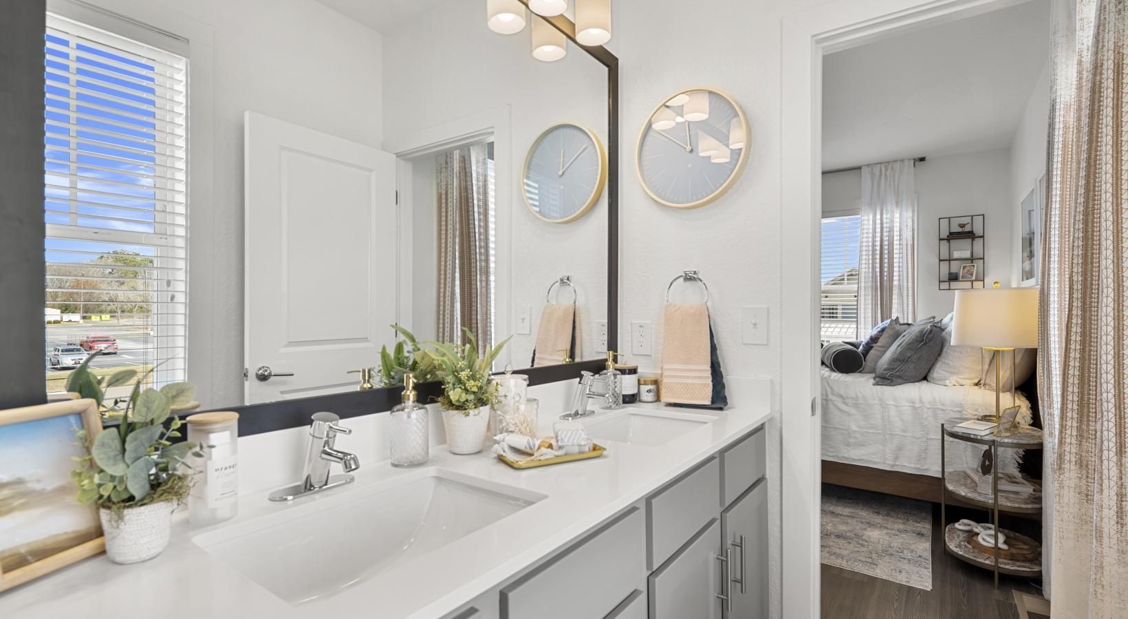 Modern bathroom vanity with dual sinks, a large mirror, and a view into a bright bedroom.
