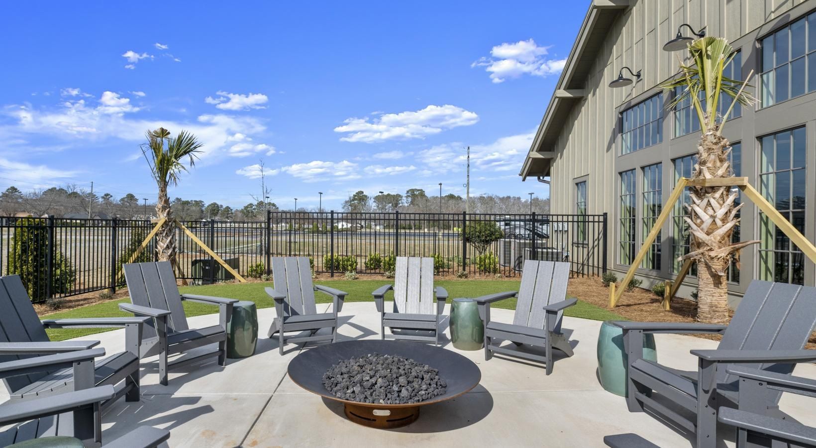 Outdoor communal patio with gray Adirondack chairs around a round fire pit beside a modern apartment building.