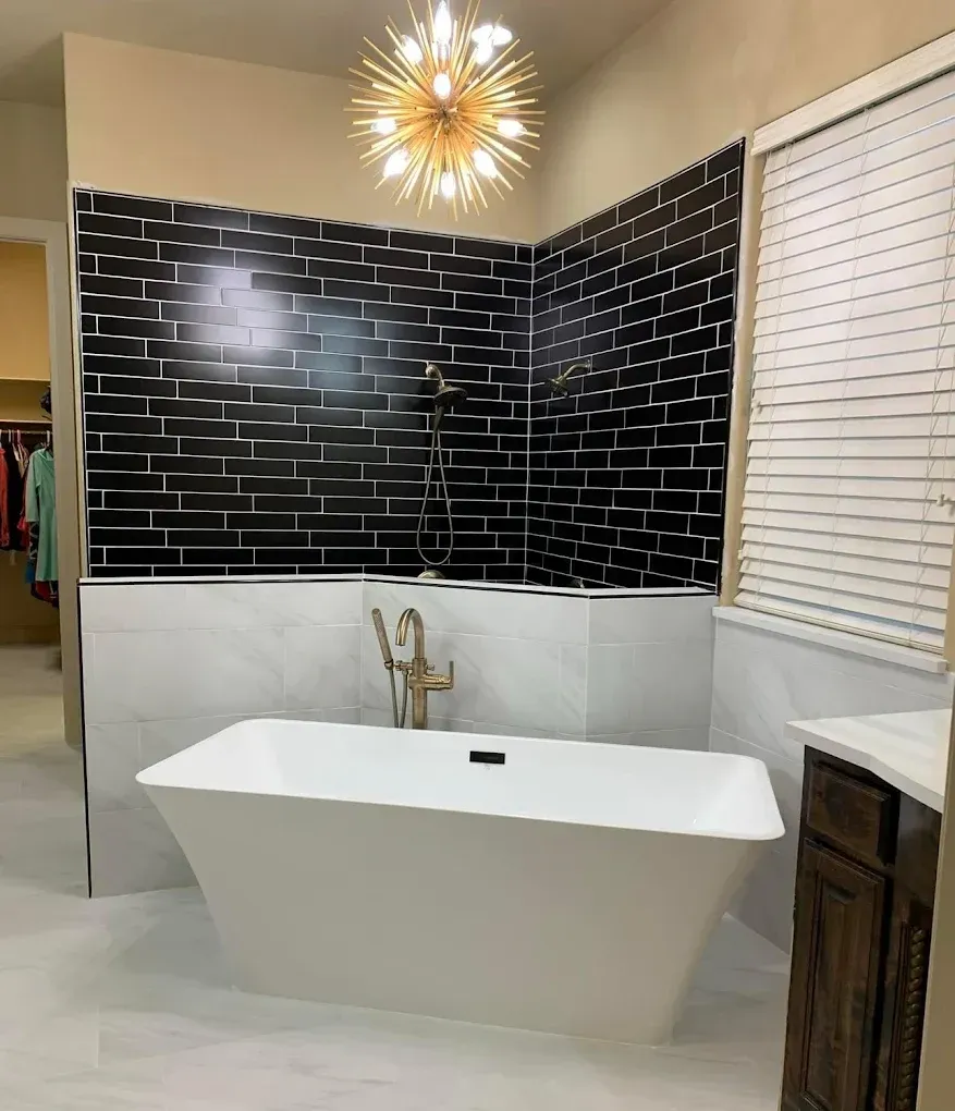 Modern bathroom with black brick-style tile, white soaking tub, gold fixtures, and a starburst chandelier.
