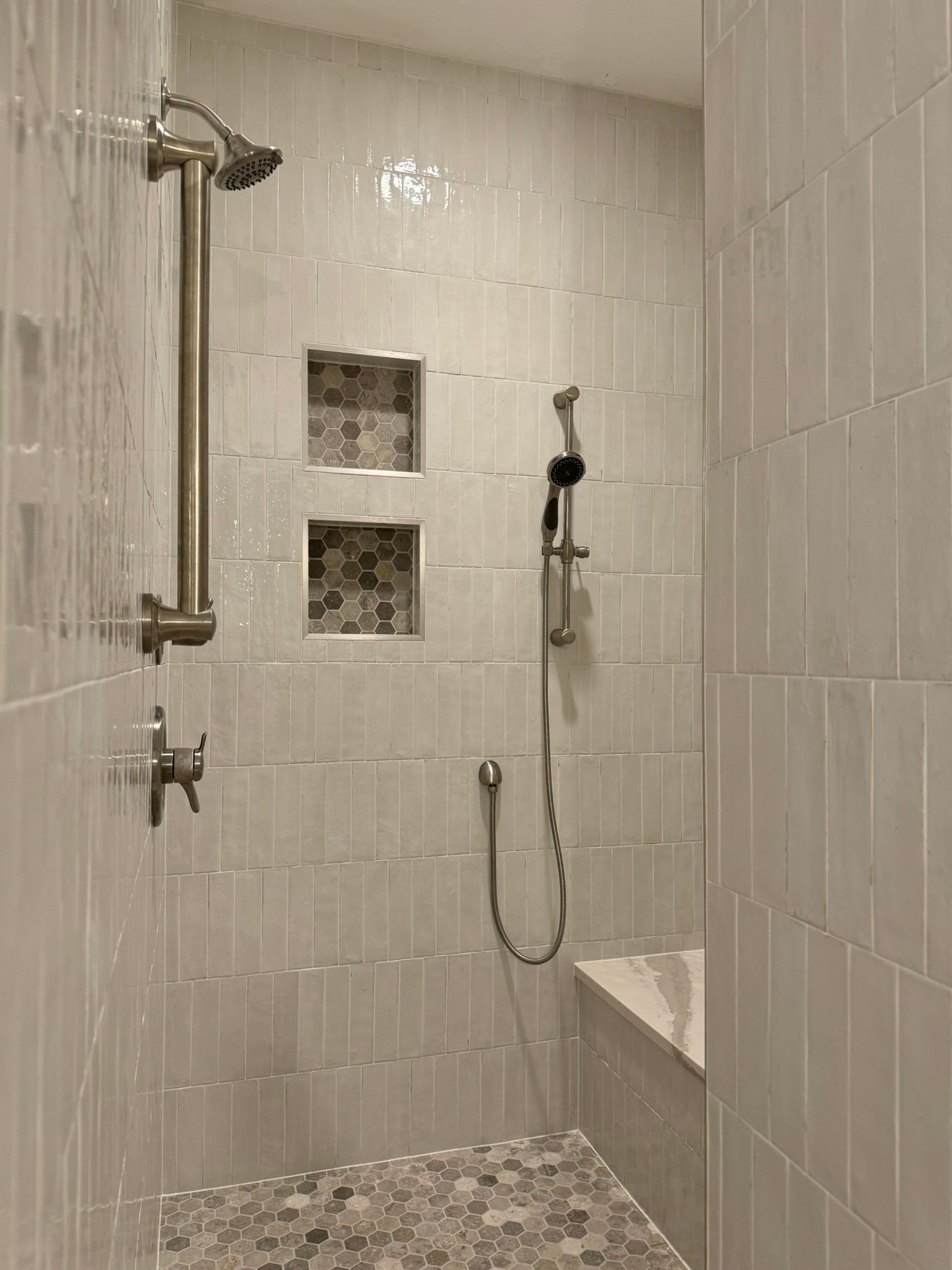Shower with light gray rectangular tiles, two recessed niches, pebble floor, and bench.