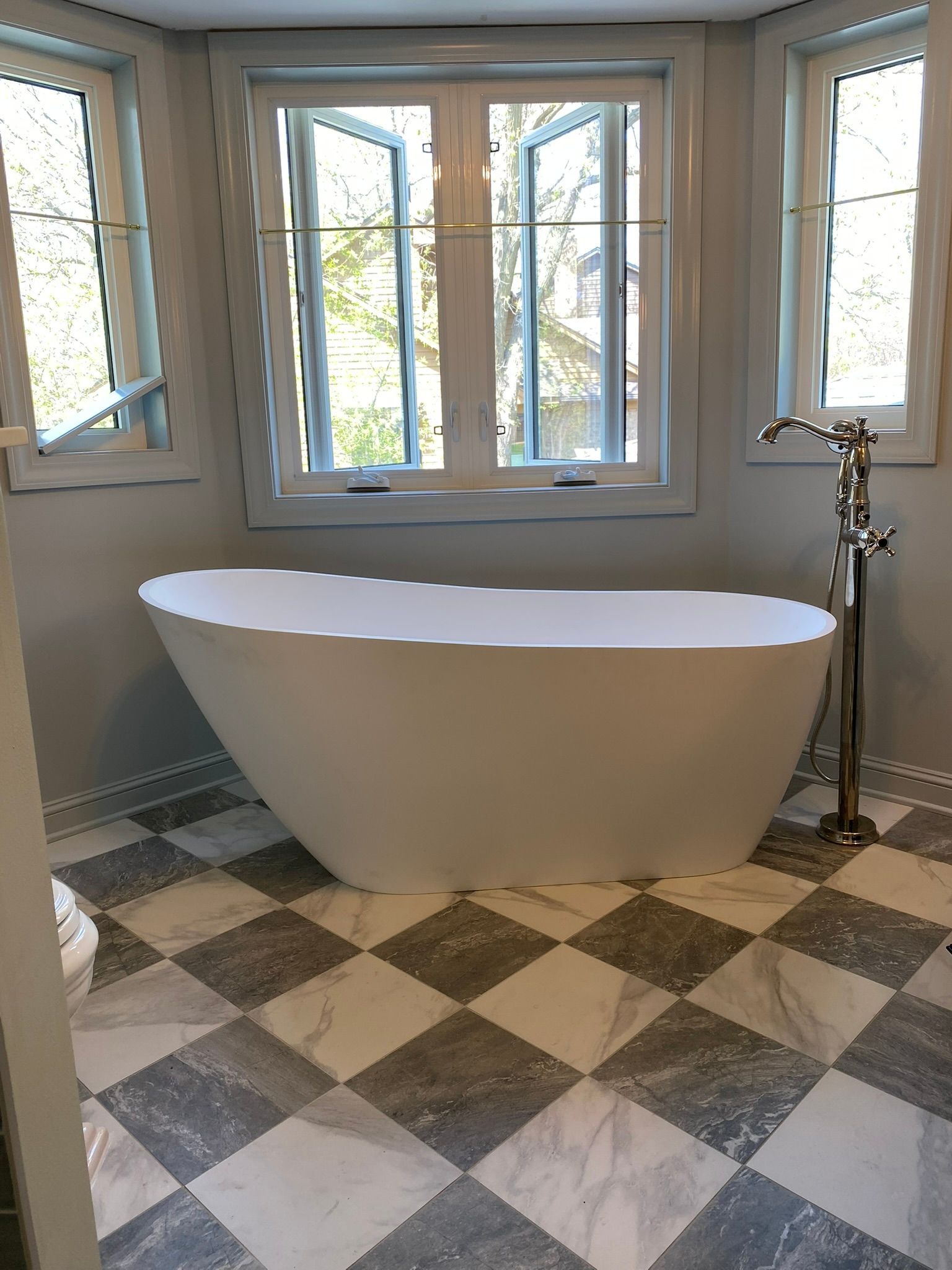 Freestanding white bathtub in a bathroom with checkered gray and white tile flooring and windows.
