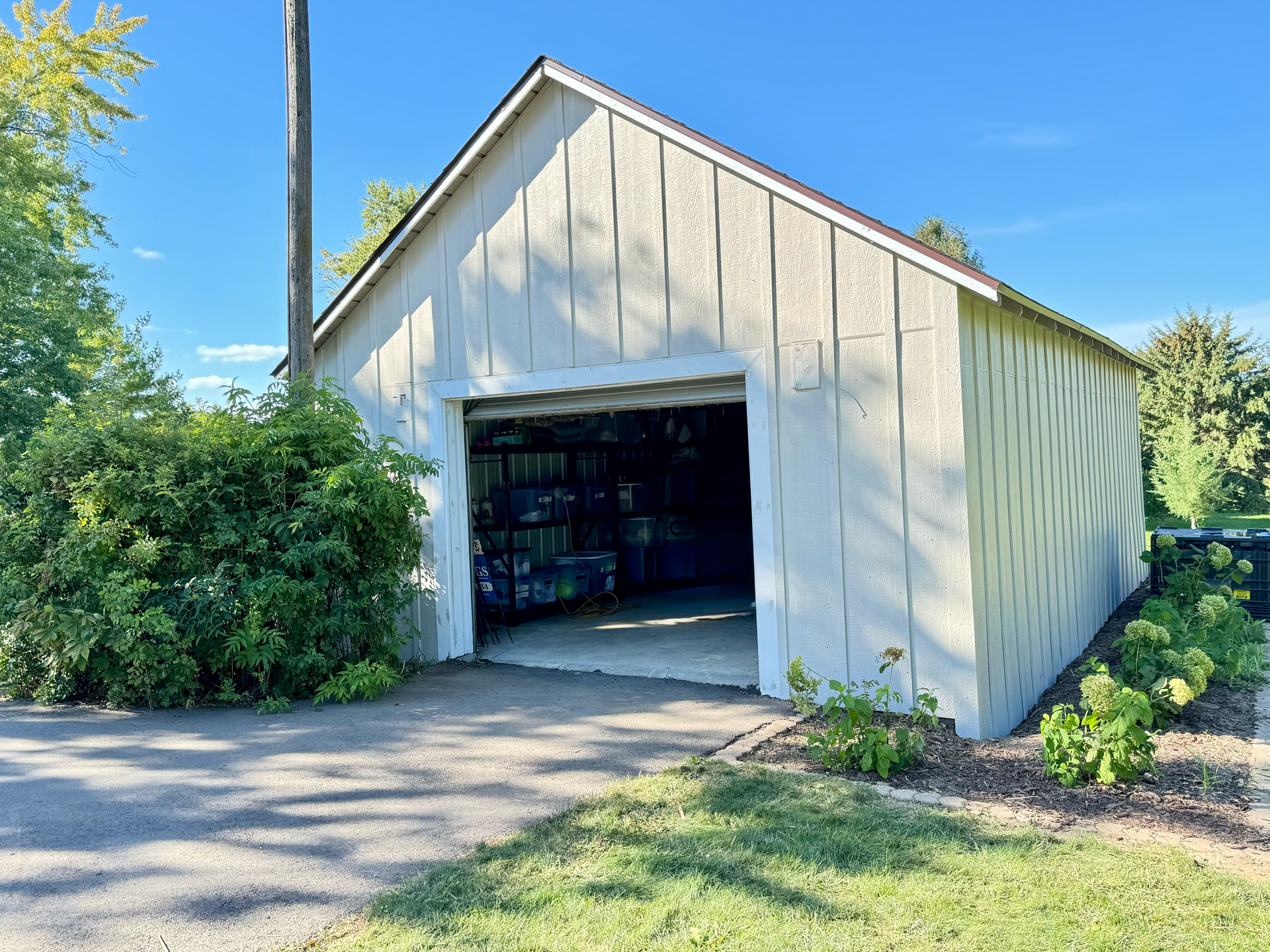 Light-colored shed with open doorway, on a paved driveway with green bushes and grass, clear sky.