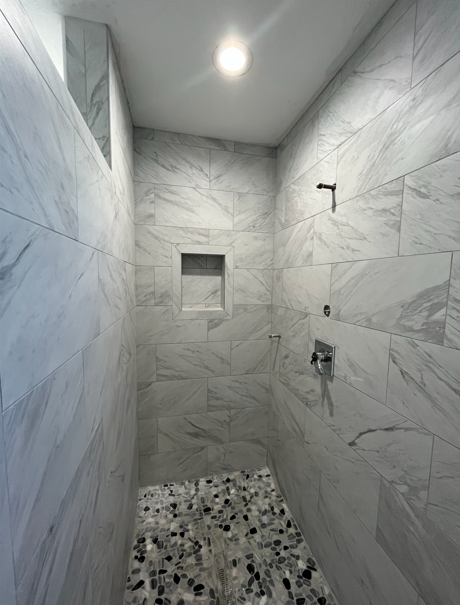 A newly tiled shower with marble-like walls, stone floor, built-in shelf, and chrome fixtures.