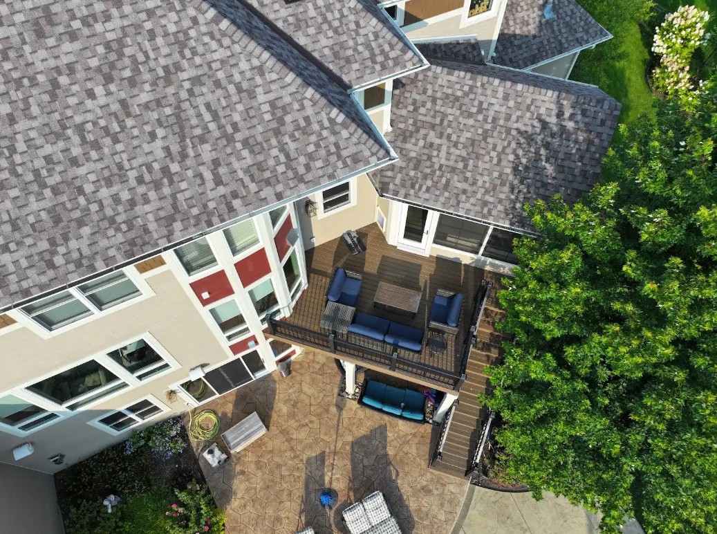 Overhead view of a house with a deck, gray roof, and surrounding trees. Brown patio, blue patio furniture.