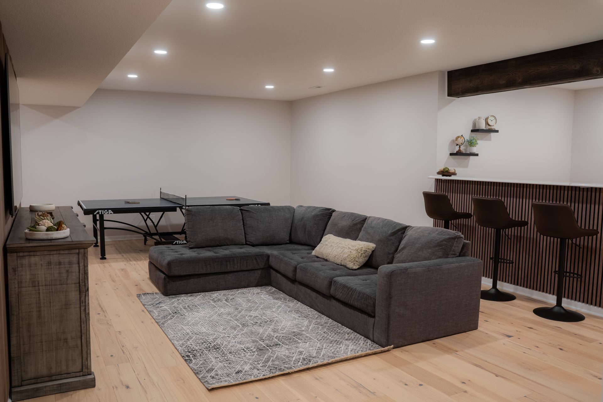 A finished basement with a sectional sofa, rug, ping pong table, and bar with stools.