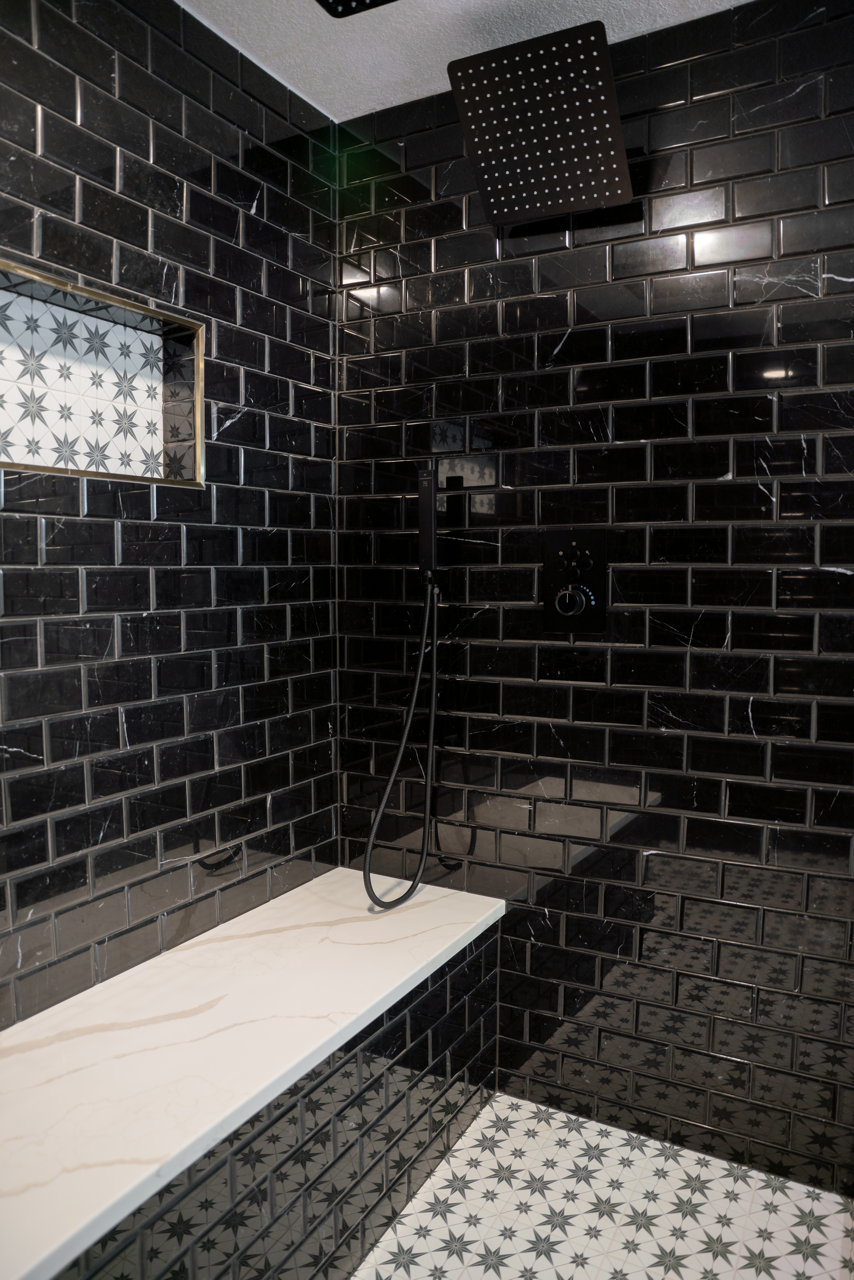 Black tiled shower with white marble bench. Black square rain shower head. Floral tile floor.