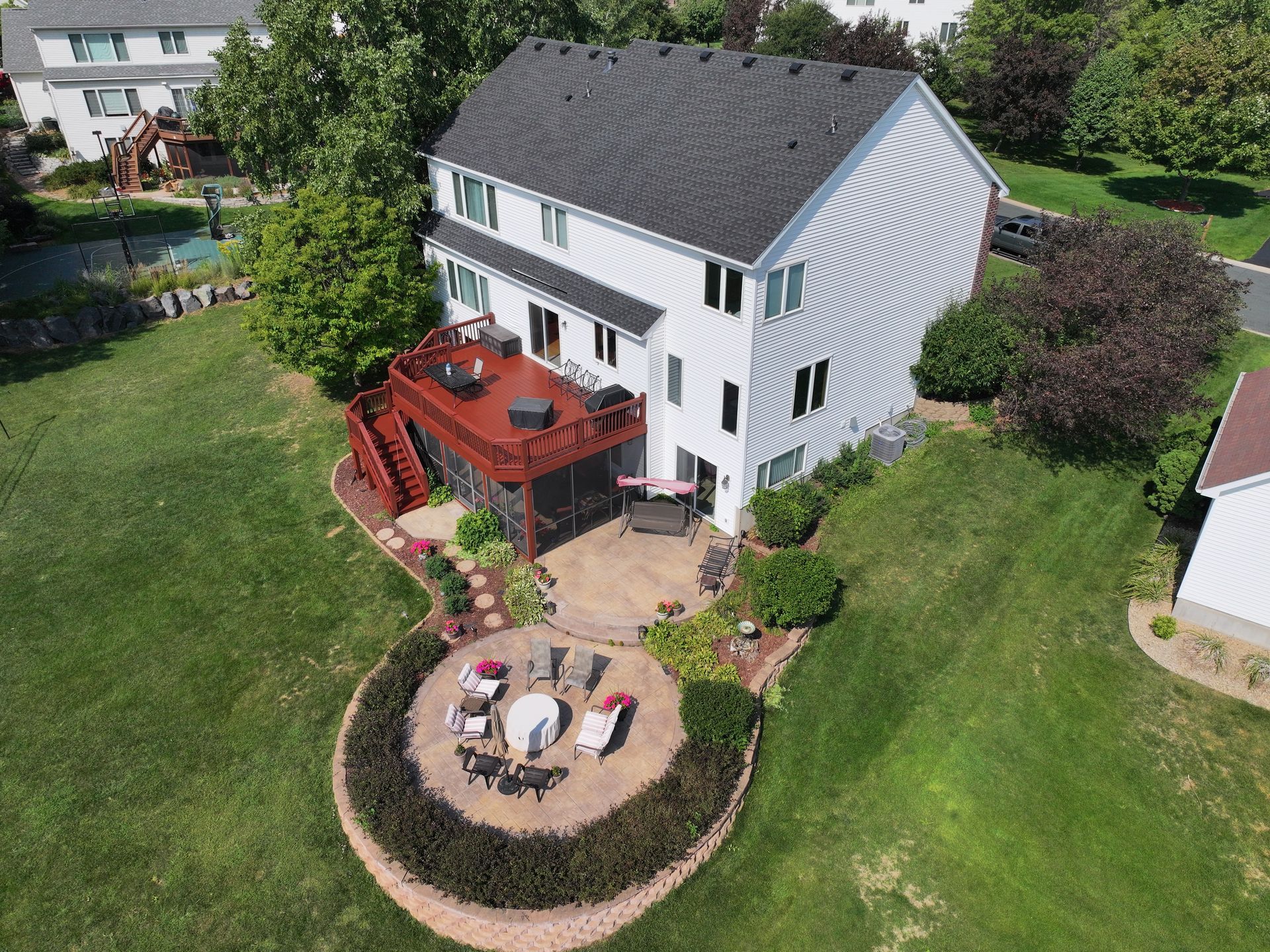 Two-story house with a wooden deck and stone patio, surrounded by green grass and landscaping.