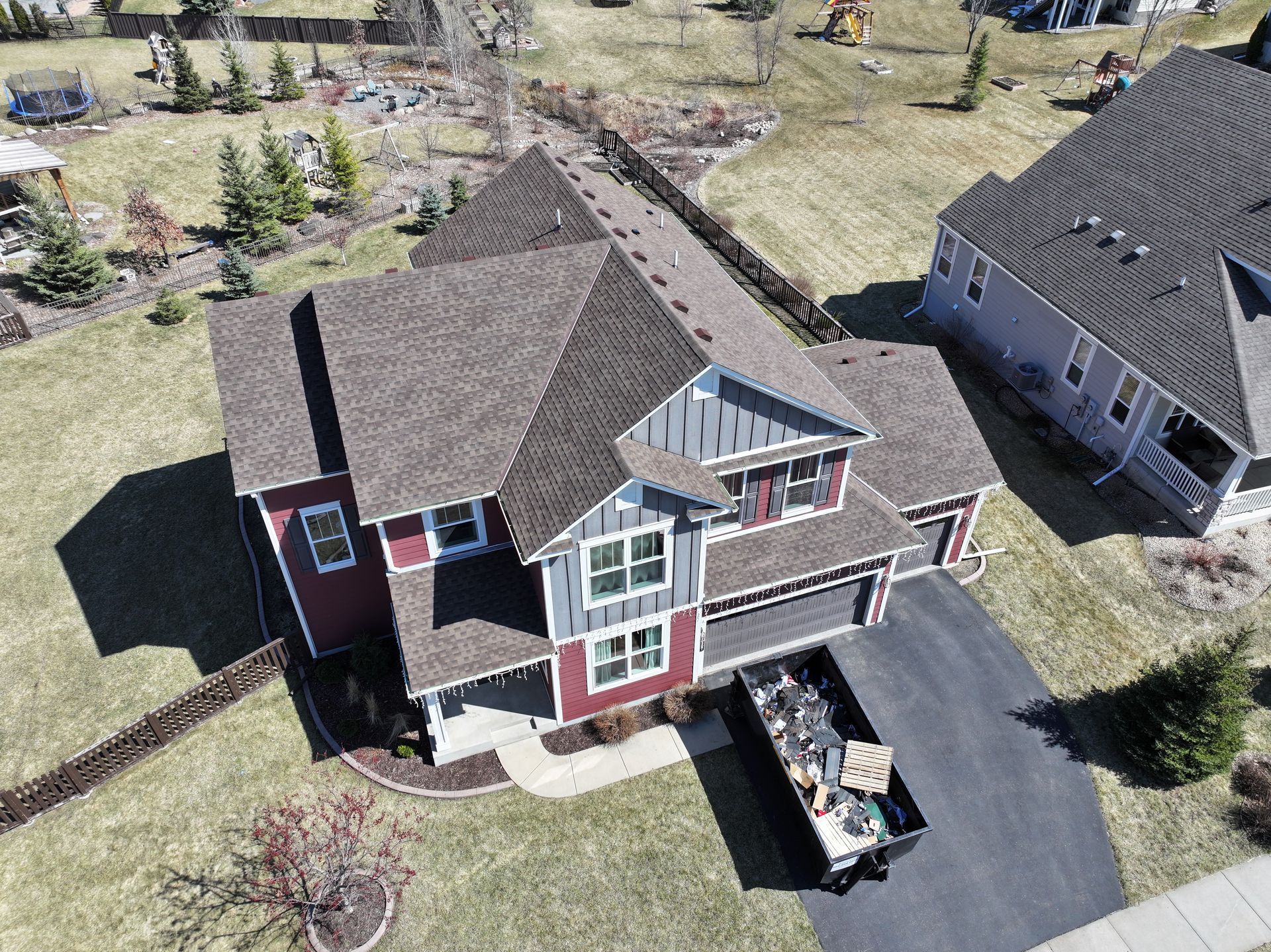 Aerial view of a two-story house with a red and grey exterior, a dark roof, and a dumpster in the driveway.