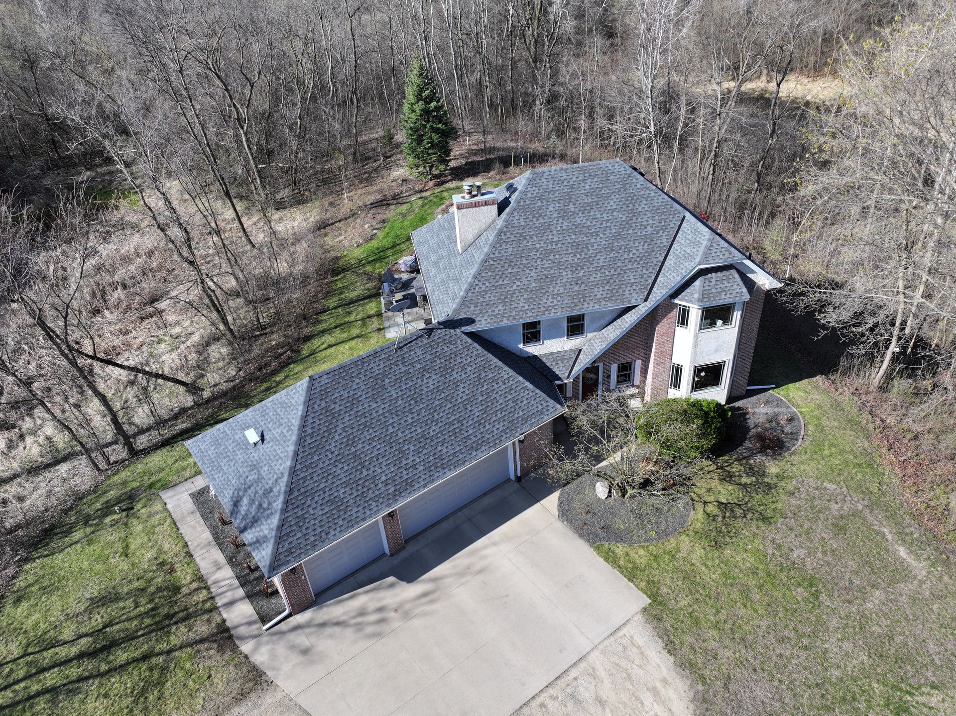 Aerial view of a gray-roofed house with a two-car garage, surrounded by bare trees and green grass.