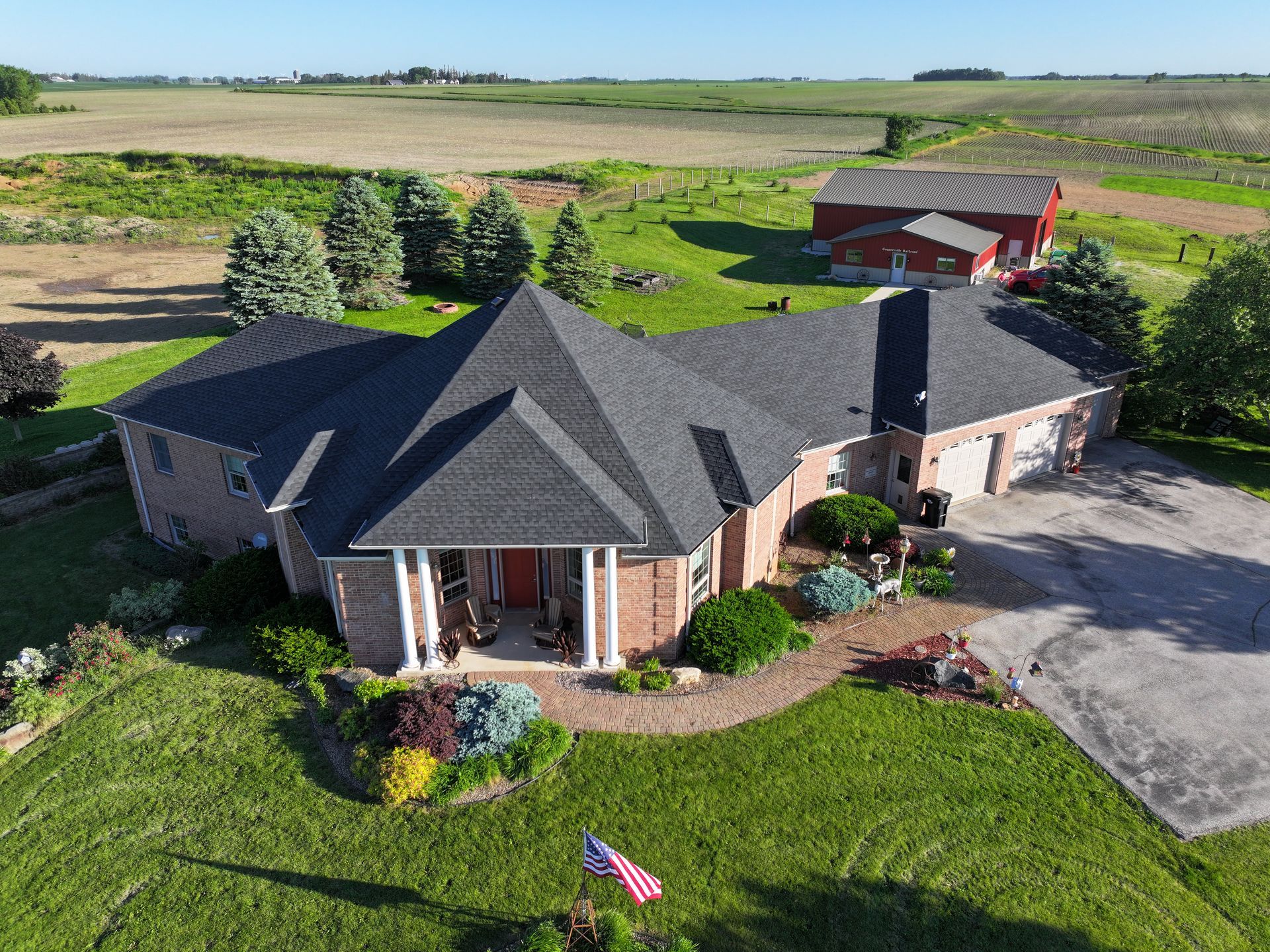 Brick house with dark roof and columns, garage, and red front door, on a grassy lot, farmland in background.